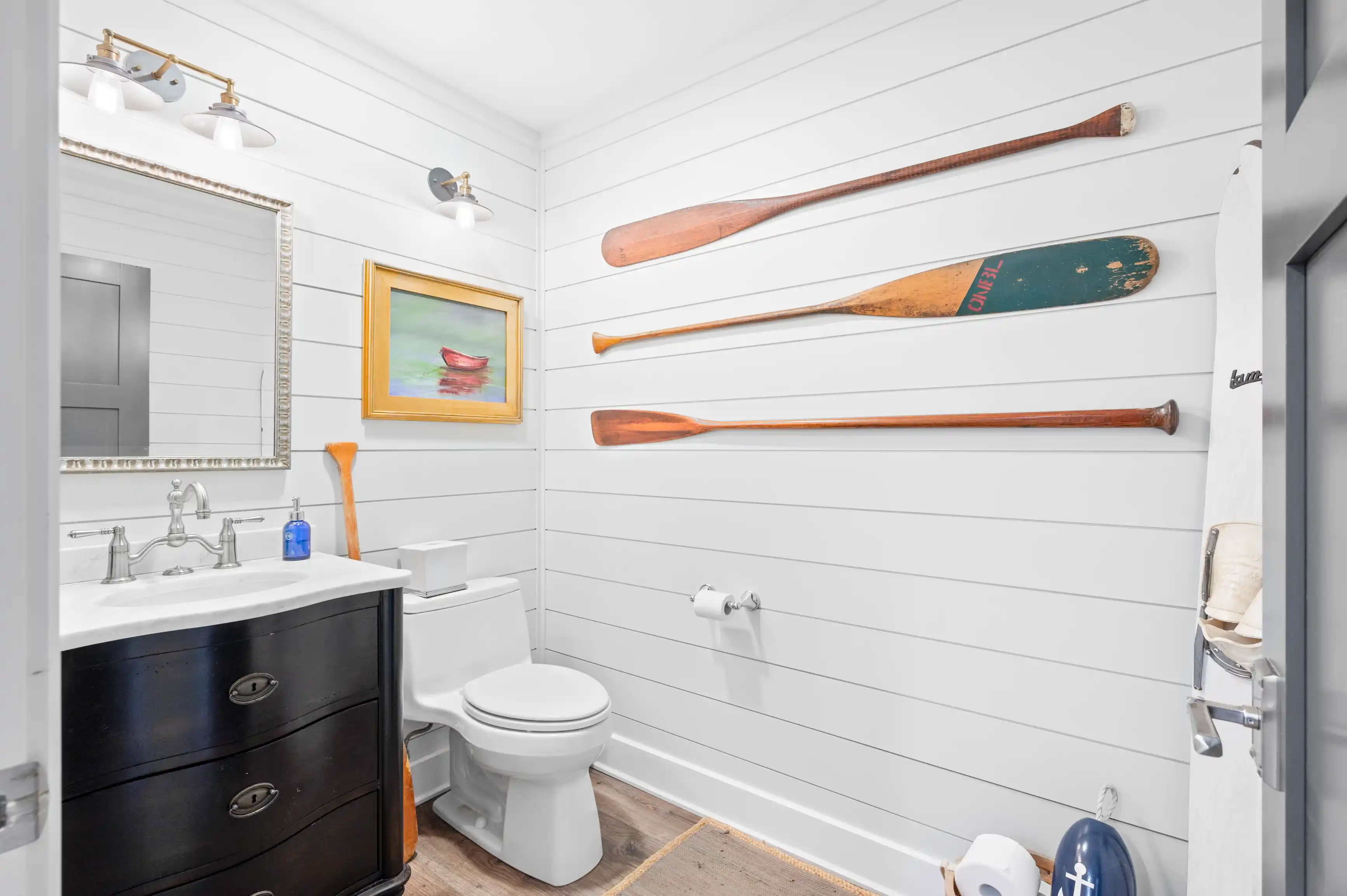 A nautical-themed bathroom with white shiplap walls, adorned with decorative oars, a framed painting of a boat, and maritime accessories.