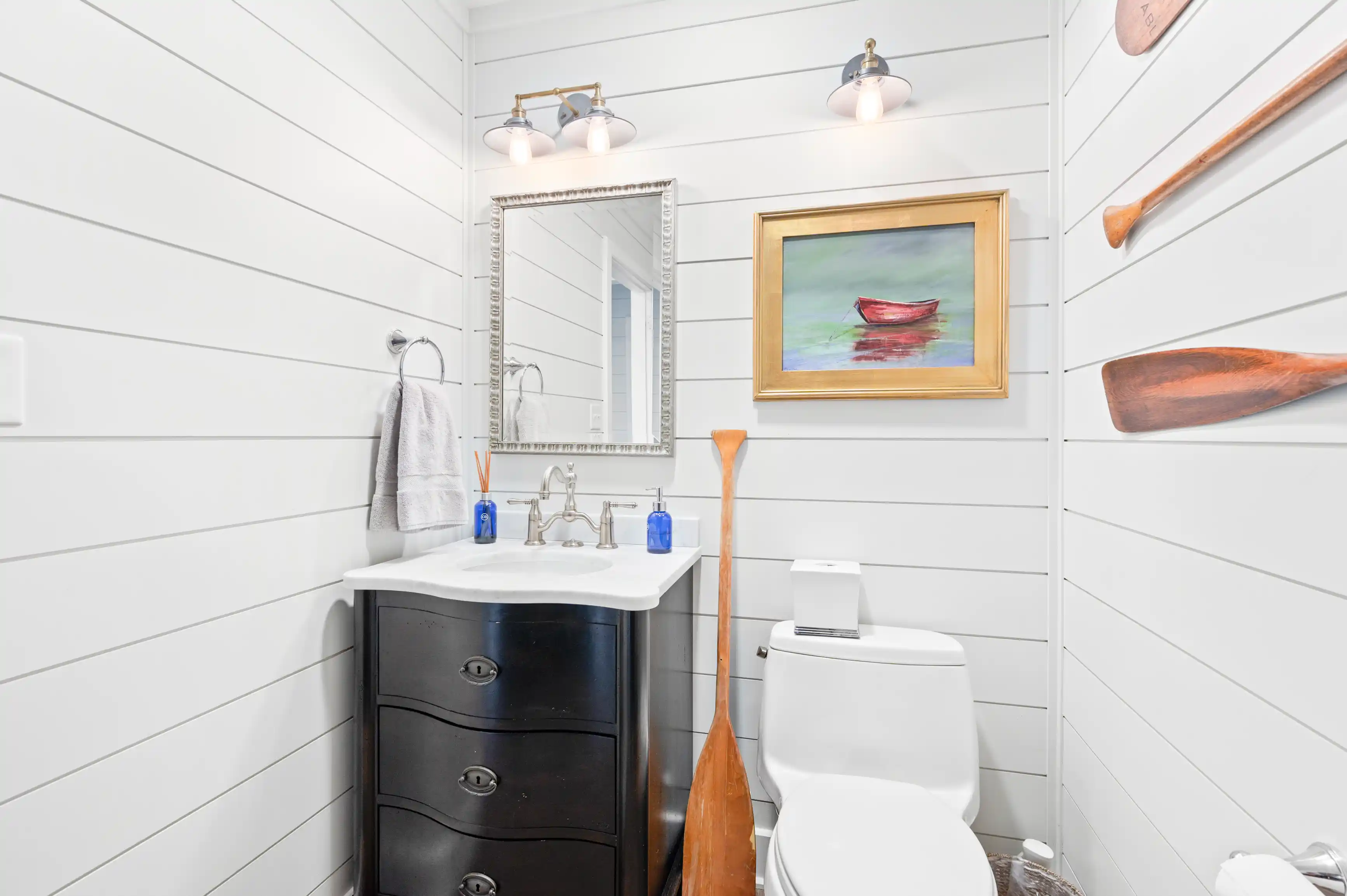 A bright, nautical-themed bathroom with white shiplap walls, a framed mirror above a sink, maritime decor including a painting of a red boat, and oars mounted on the wall.