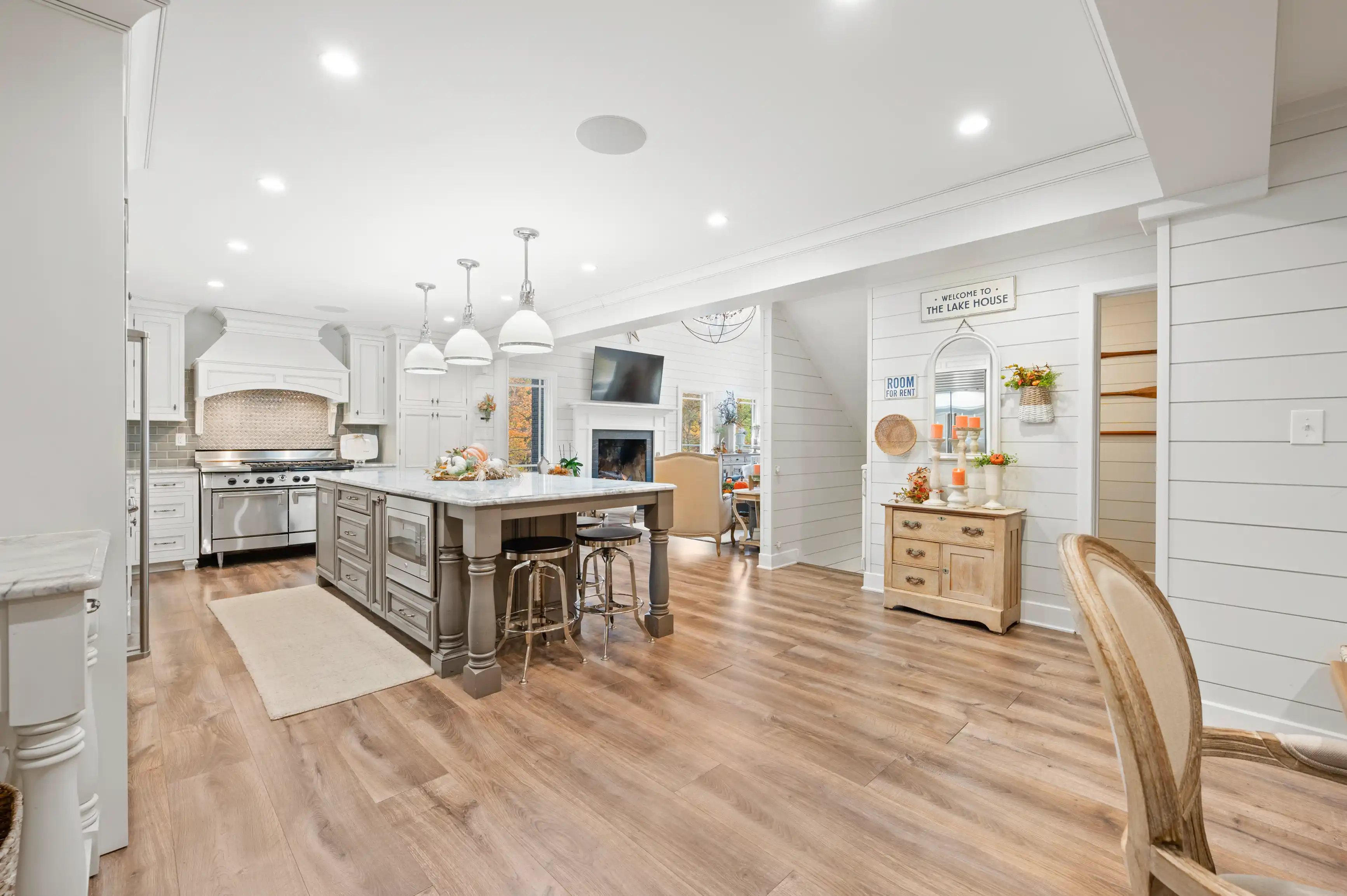 Bright and spacious kitchen interior with white cabinetry, a central island with stools, wooden floors, and an adjoining cozy dining area with fireplace.