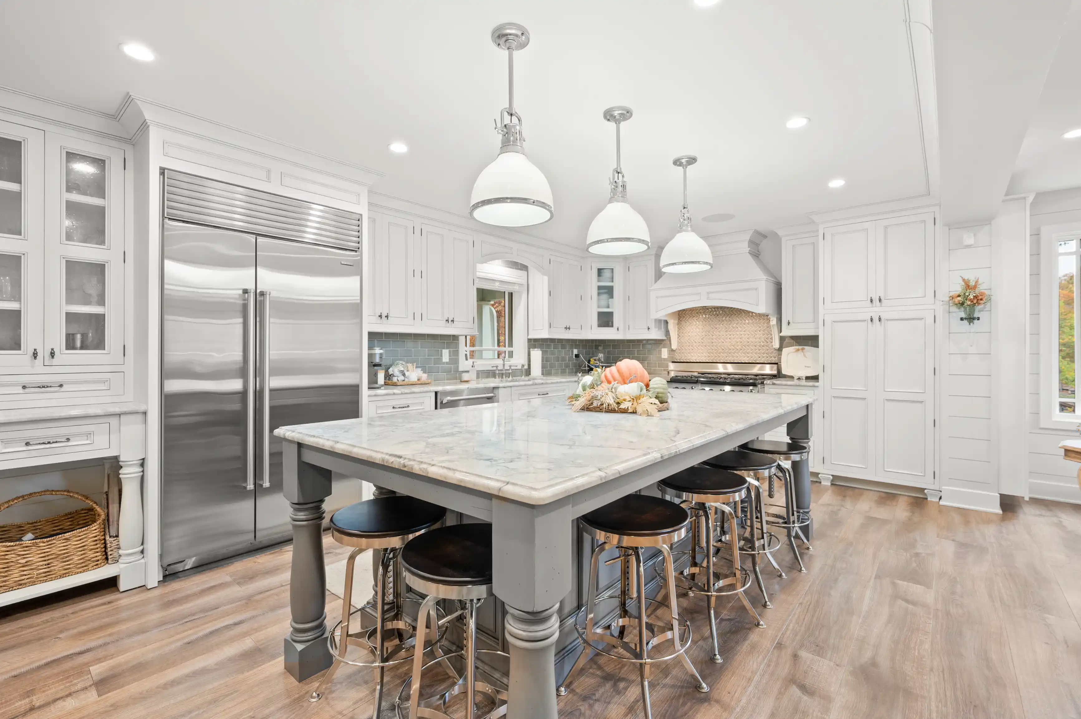 Bright contemporary kitchen interior with white cabinets, marble countertop island, bar stools, stainless steel appliances, and pendant lighting.