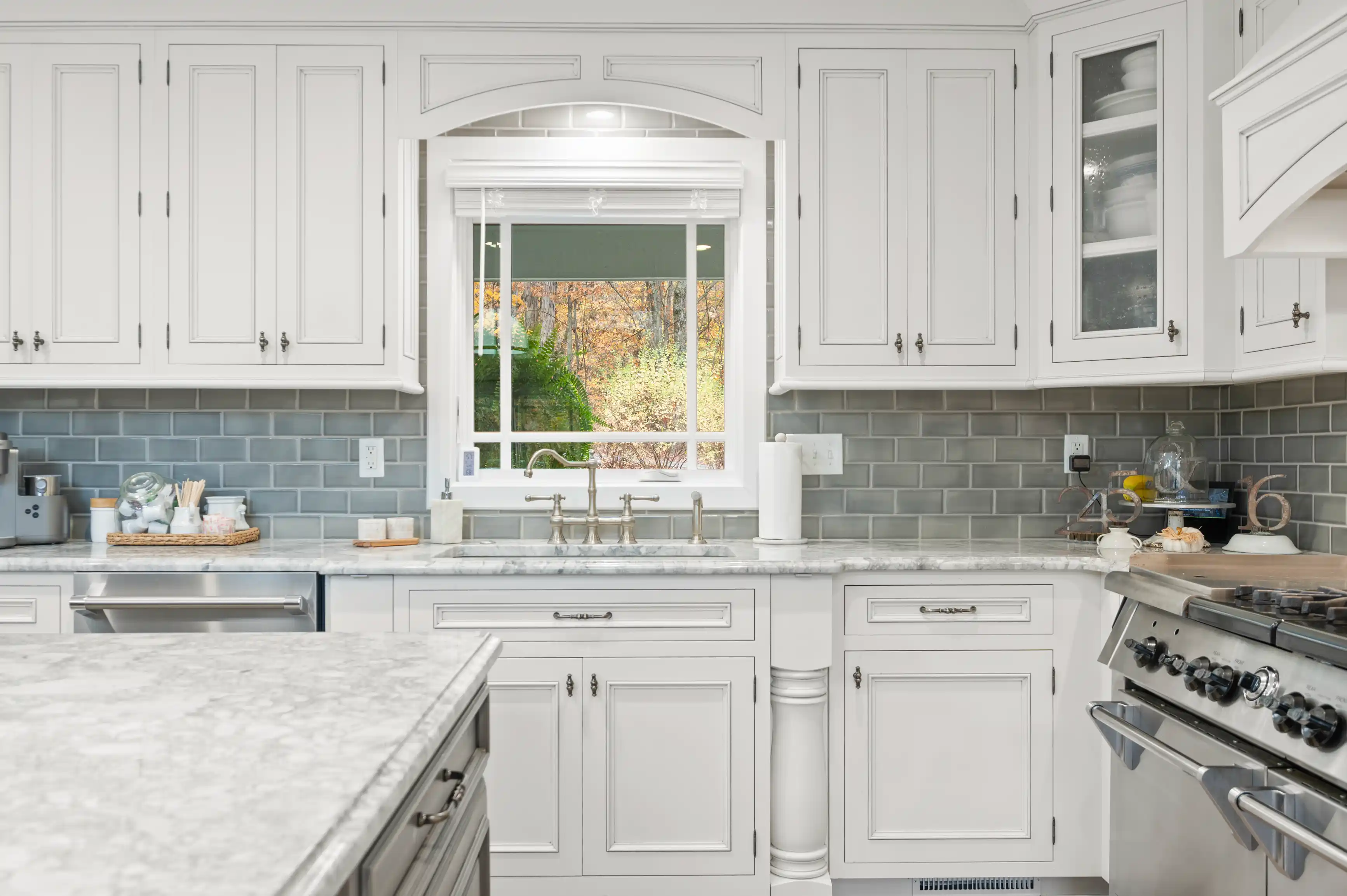 Elegant kitchen interior with white cabinetry, marble countertops, and blue subway tile backsplash, featuring a window with a view of autumn foliage.