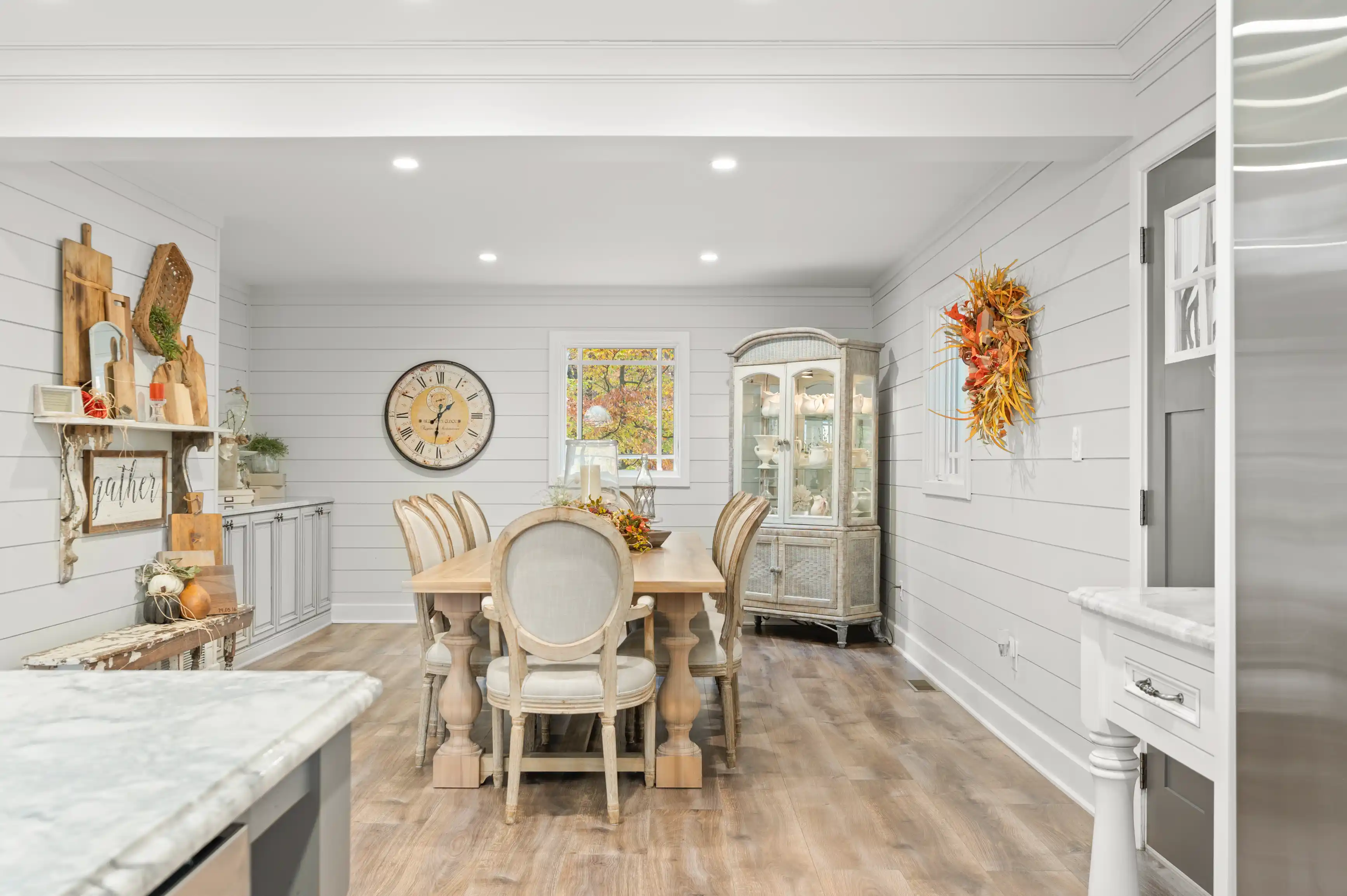 Bright and inviting dining room with a rustic wooden table and chairs, shiplap walls, autumn decor accents, and a vintage-style cabinet.
