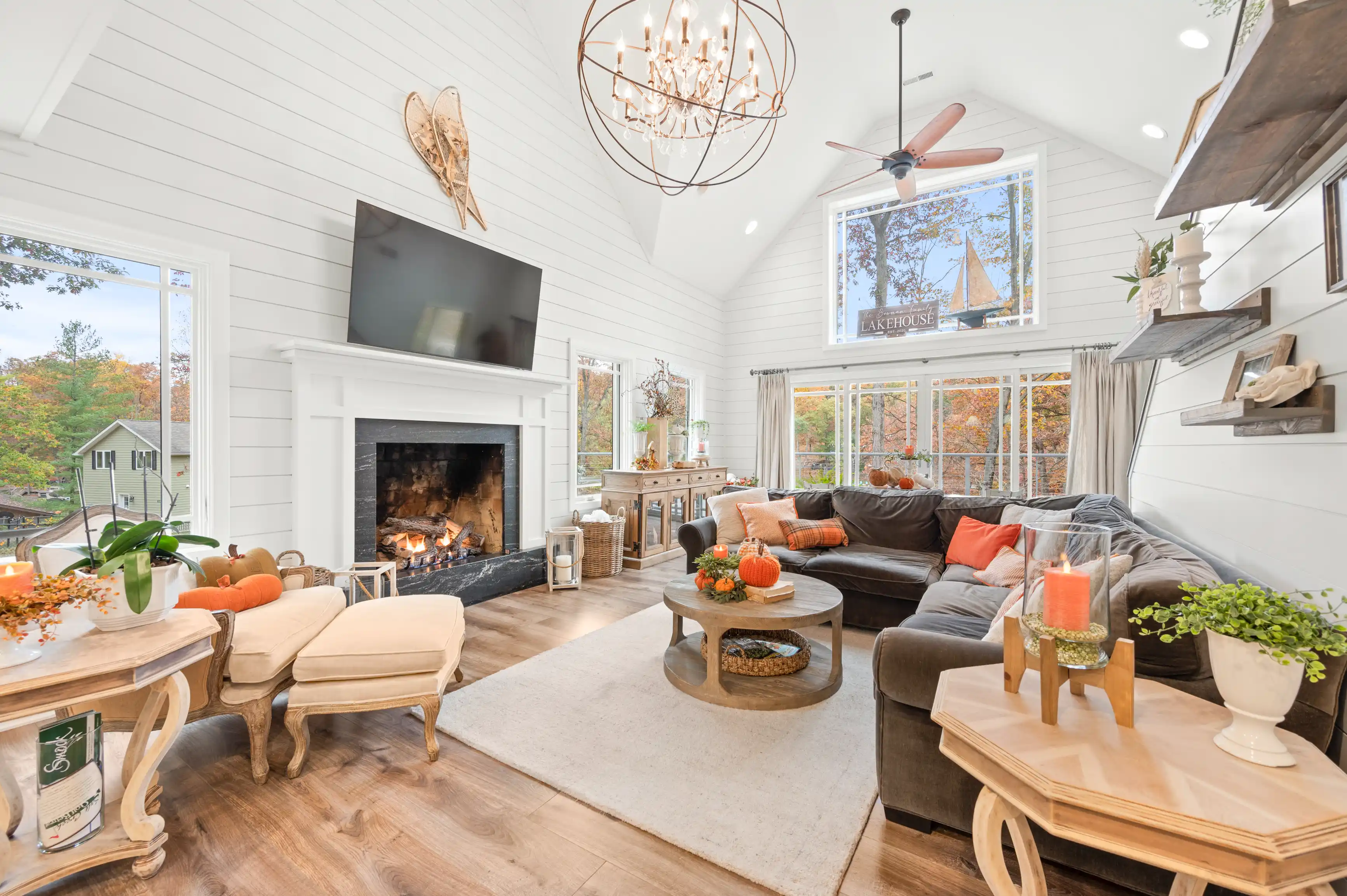 Bright and cozy living room interior with a lit fireplace, comfortable furniture, and large windows overlooking autumn foliage.
