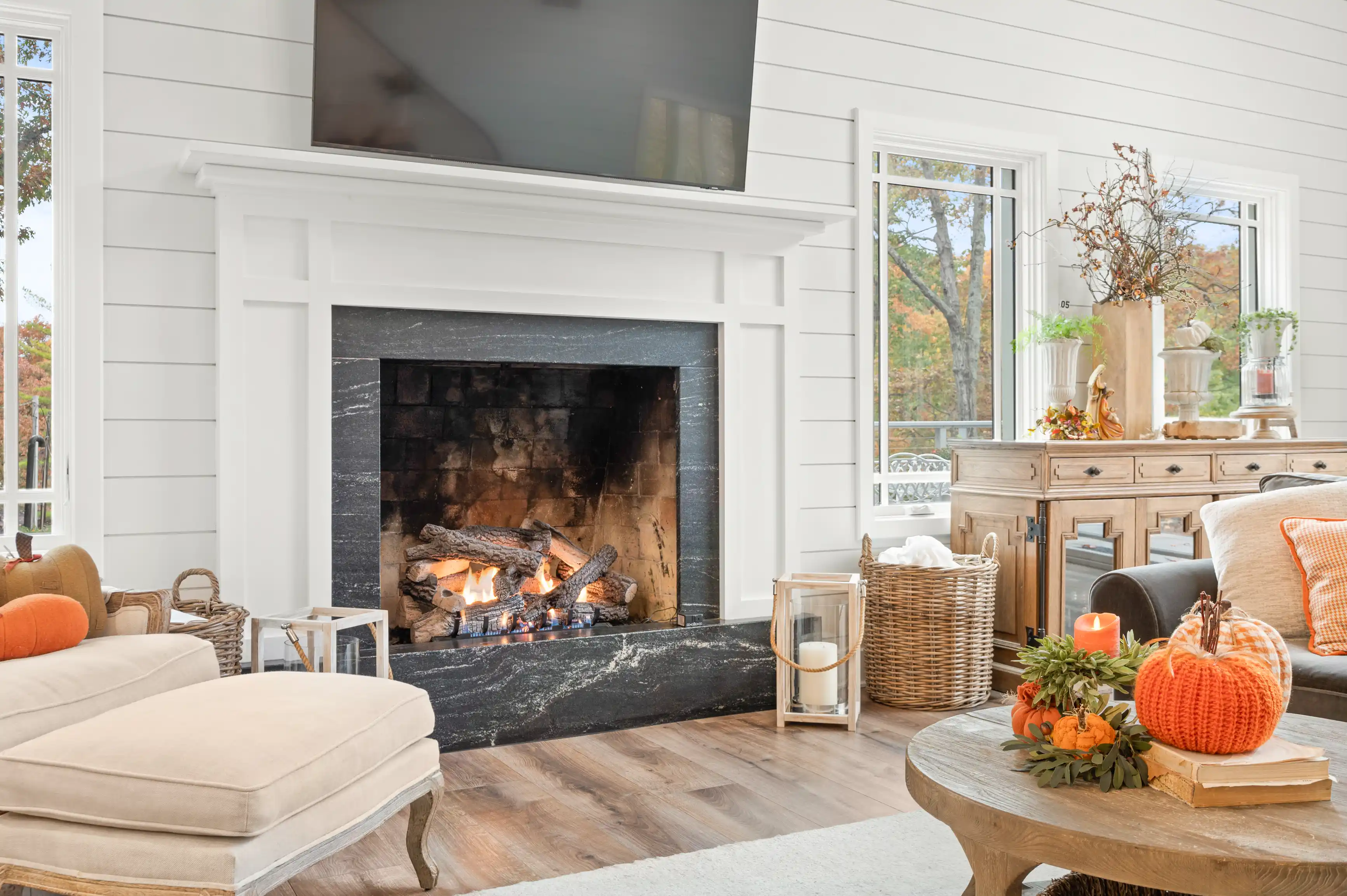 Cozy living room with a lit fireplace, a beige sofa with orange pillows, wooden furniture, autumn decorations, and a view of trees through a window.
