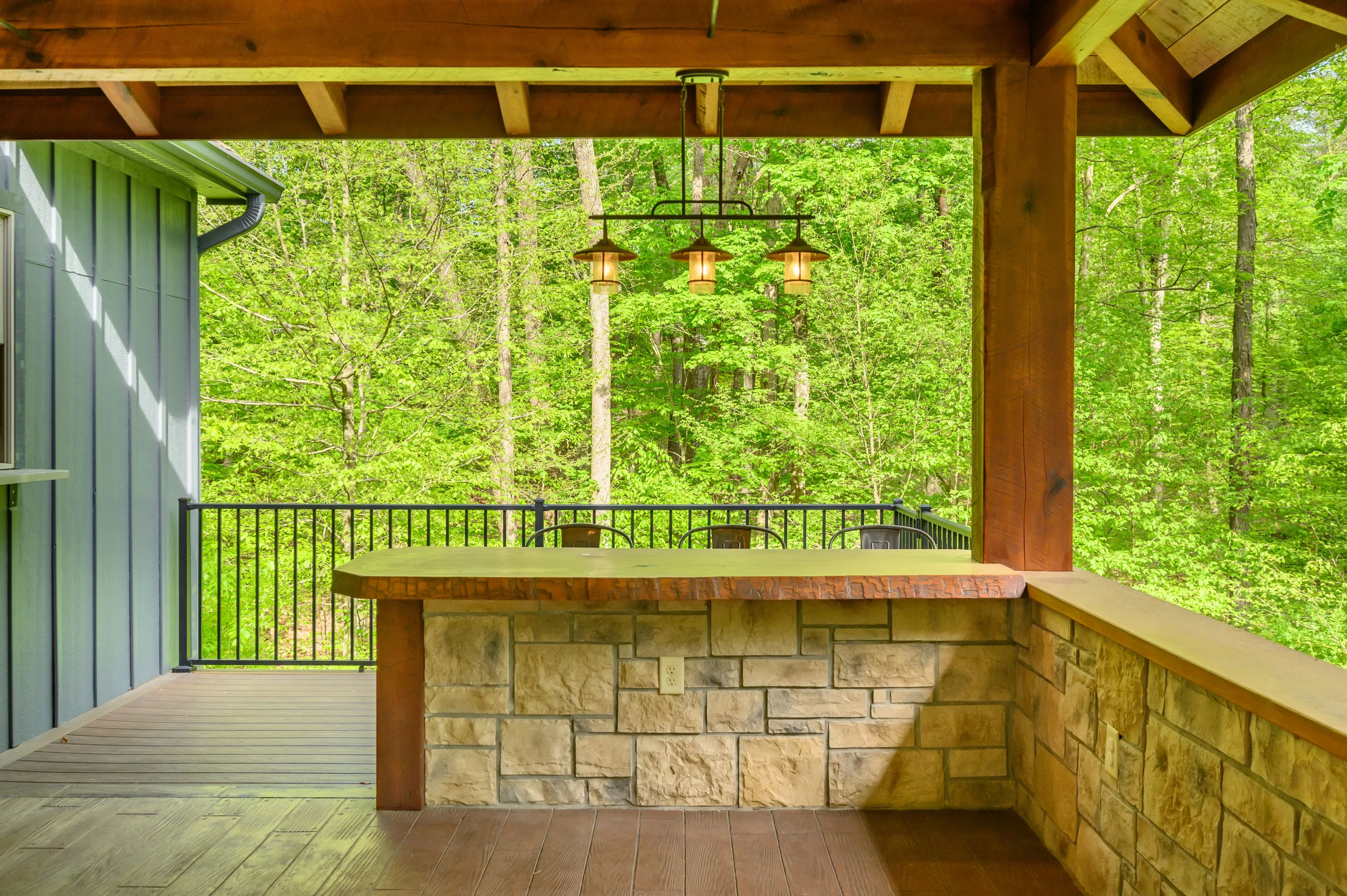 Covered wooden deck with a stone bar counter, iron railing, and forest view.