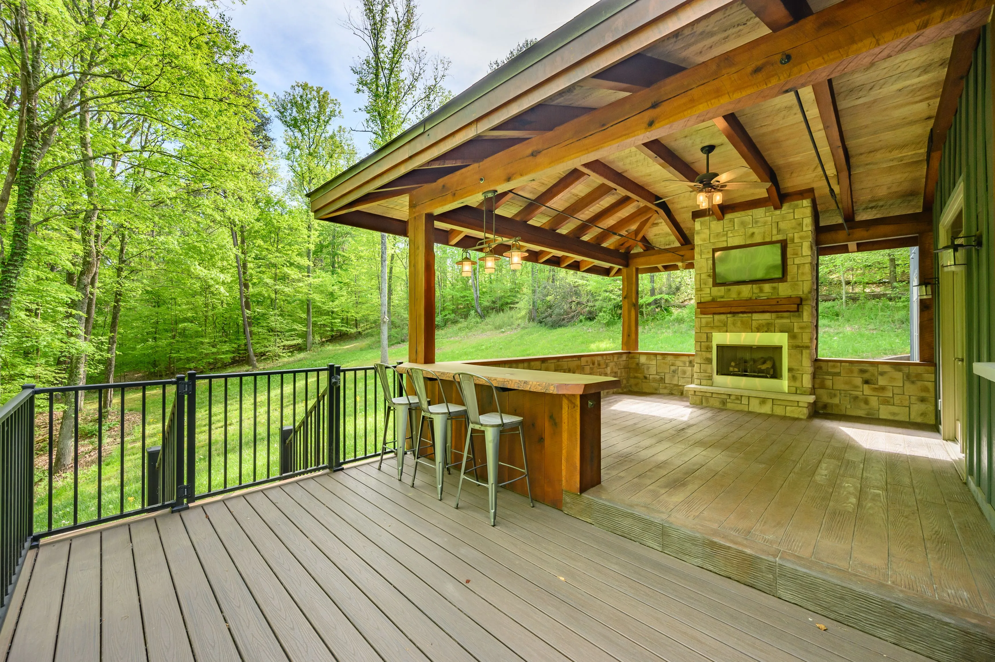 Spacious covered outdoor patio with a wooden bar, stools, fireplace, and a view of lush greenery.