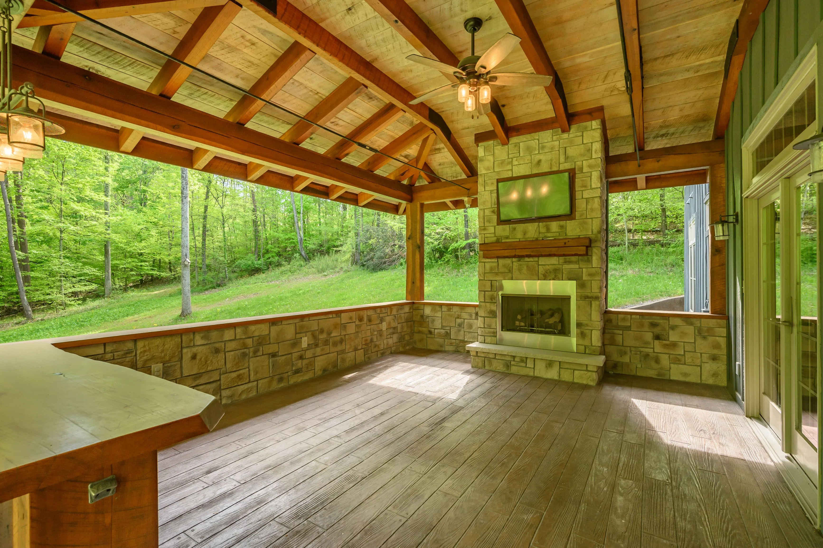 An empty sunroom with wood flooring, stone fireplace, and large windows overlooking a lush green forest.