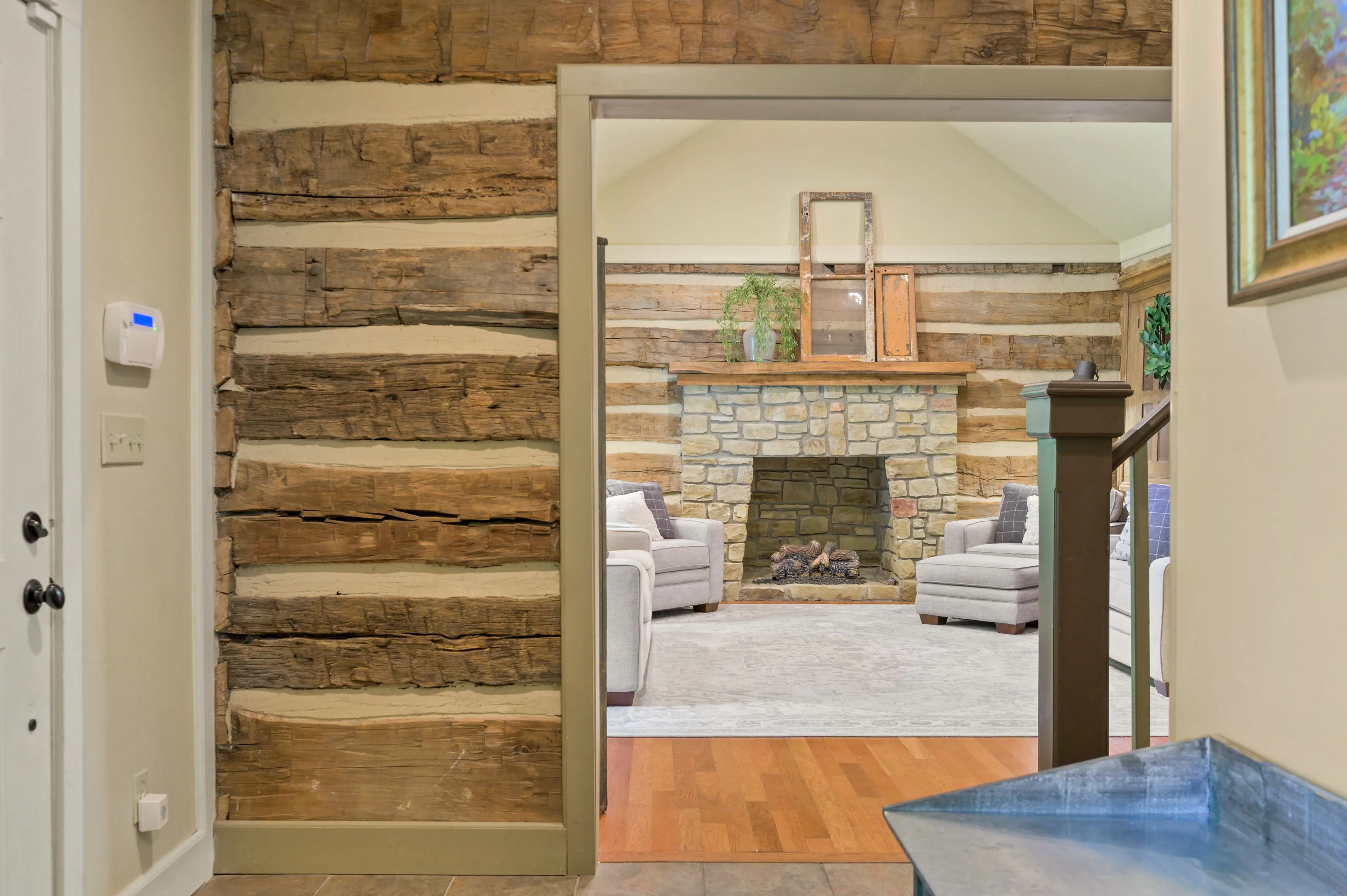 View from a hallway with rustic log cabin walls into a cozy living room with a stone fireplace, comfortable sofas, and a decorative mirror above the mantelpiece.