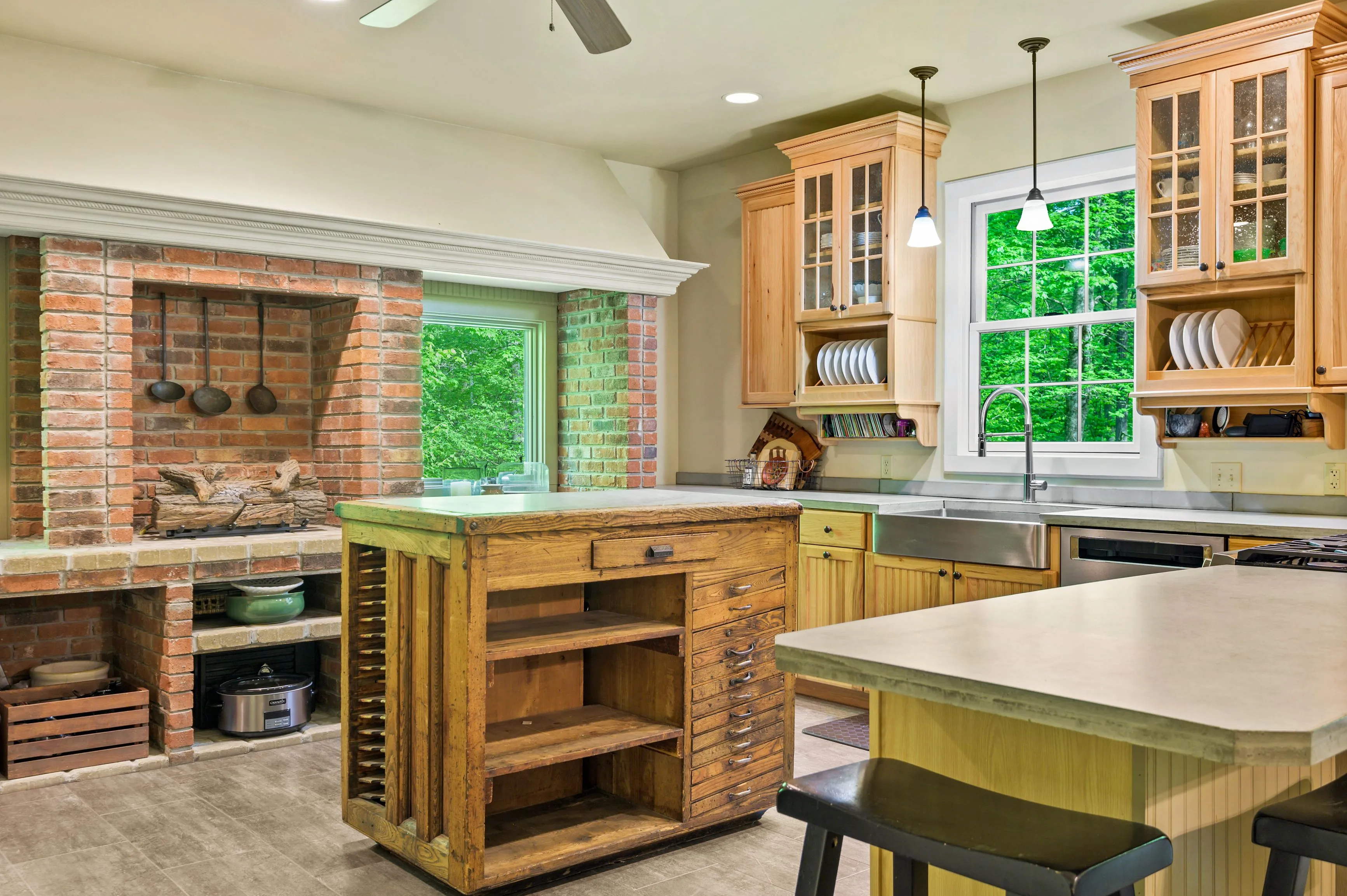 Spacious kitchen interior with wooden cabinets, large island, brick fireplace, and a view of greenery outside the window.