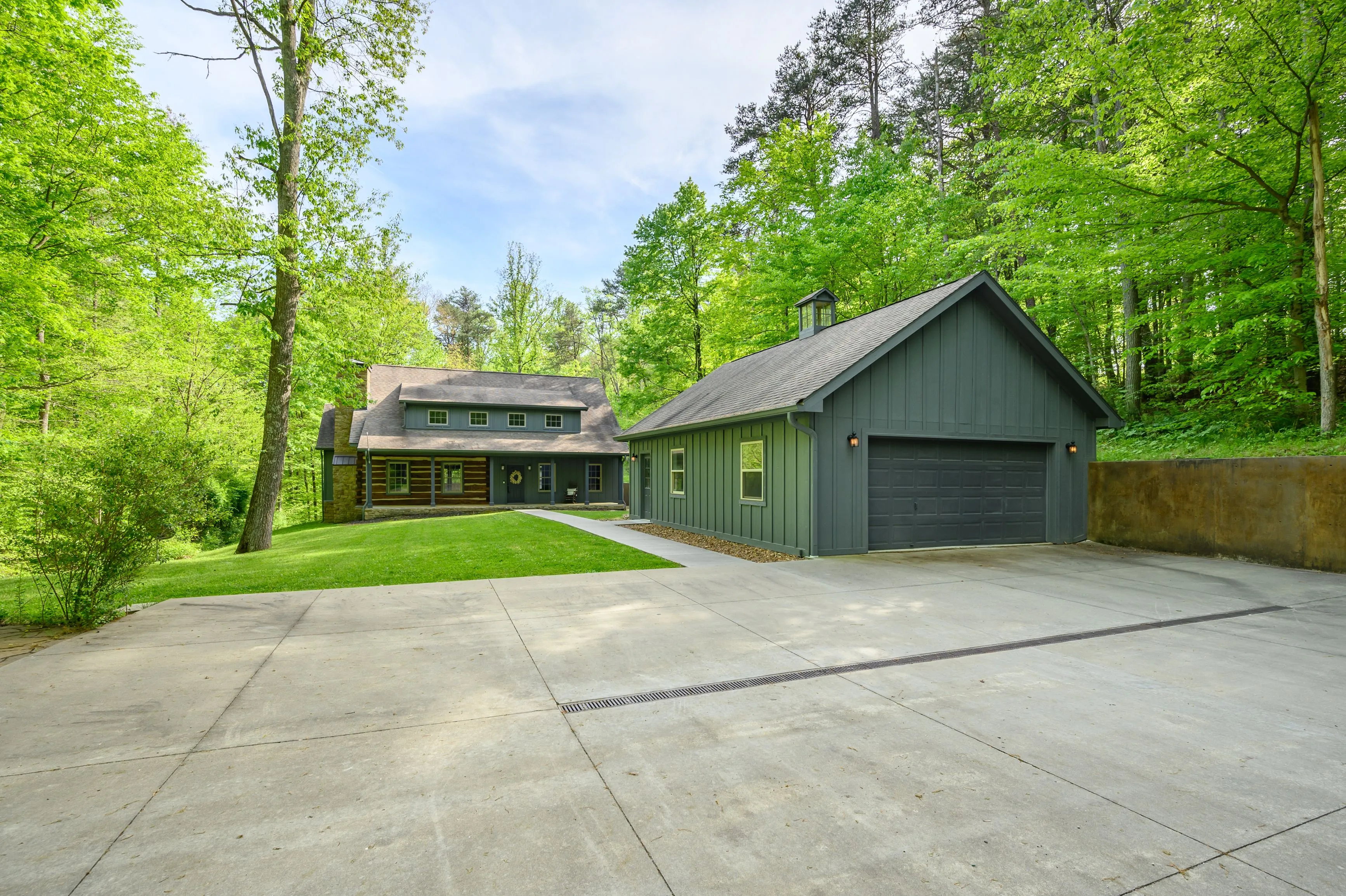 A modern two-story house with a gray exterior and attached garage surrounded by lush green trees with a concrete driveway in the foreground.
