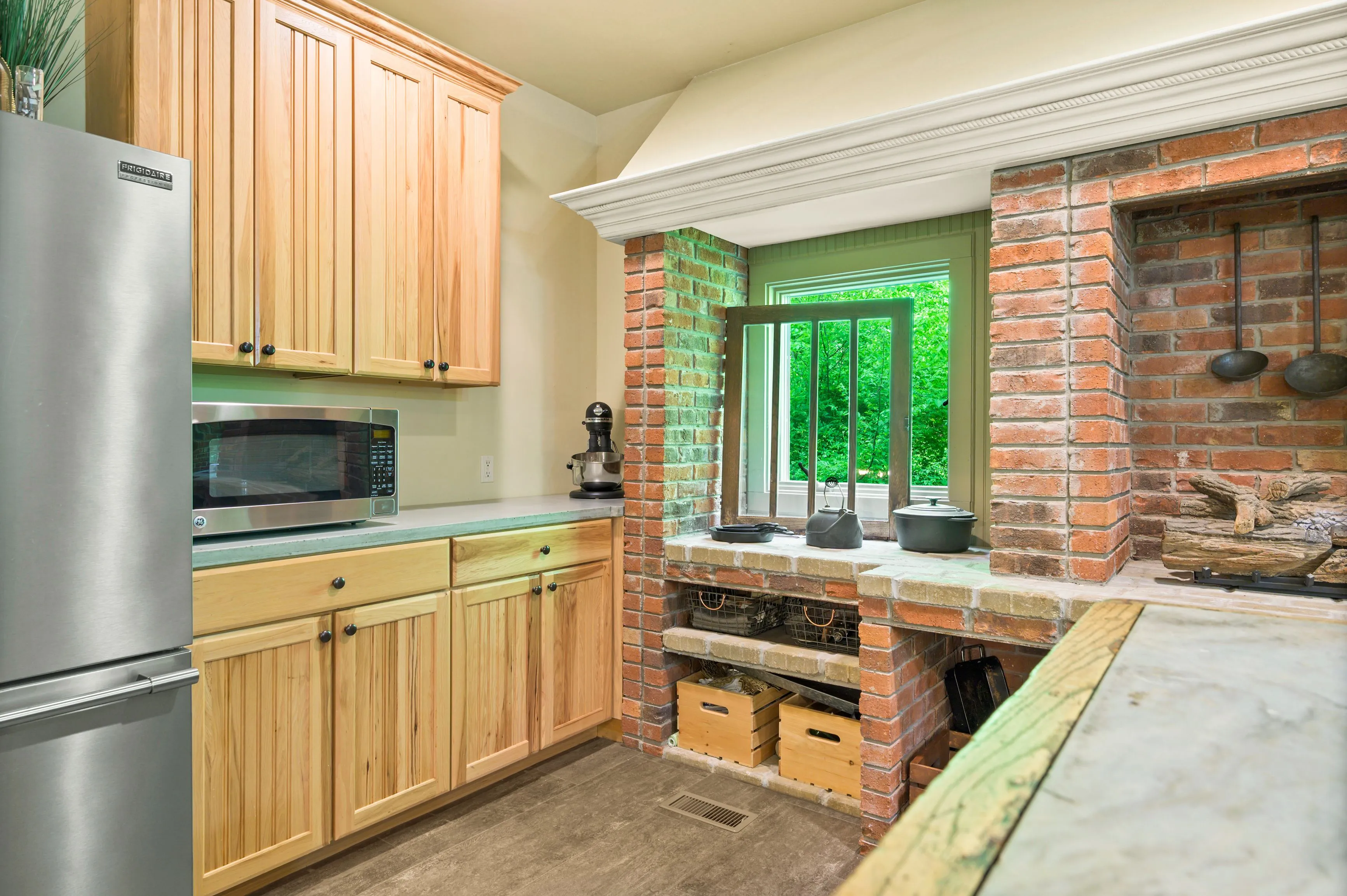 Modern kitchen interior with wooden cabinets and brick fireplace, stainless steel refrigerator and microwave, overlooking greenery through the window.