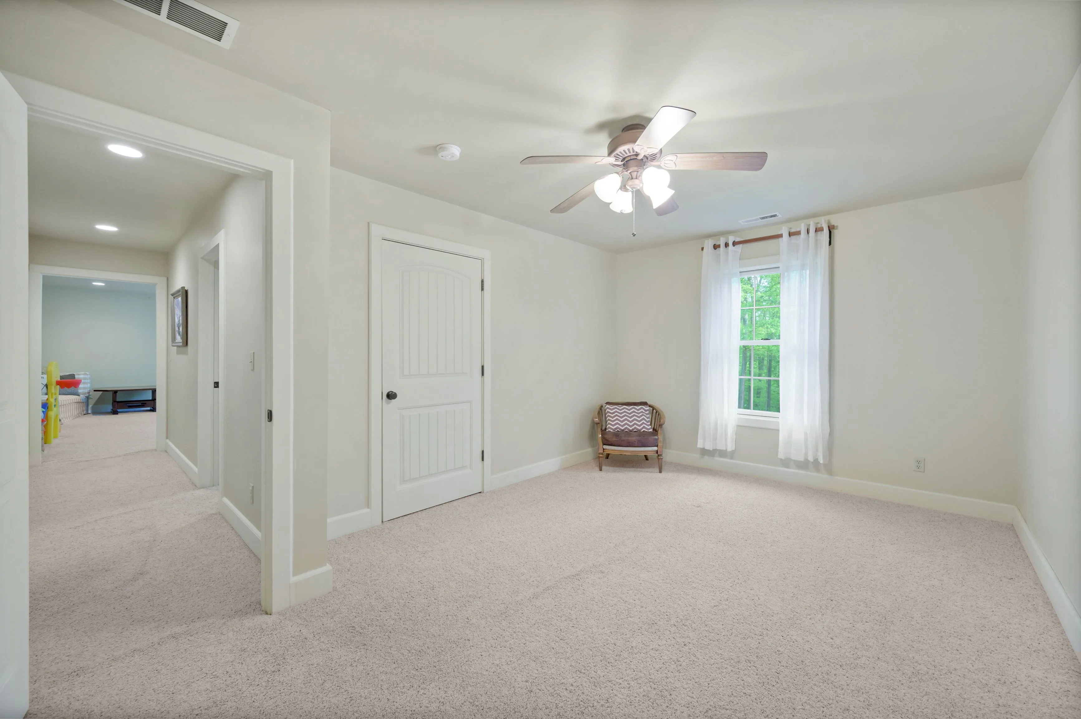 Empty room with beige carpeting, white walls, ceiling fan, a single chair by the window, and an open door showing another room in the background.