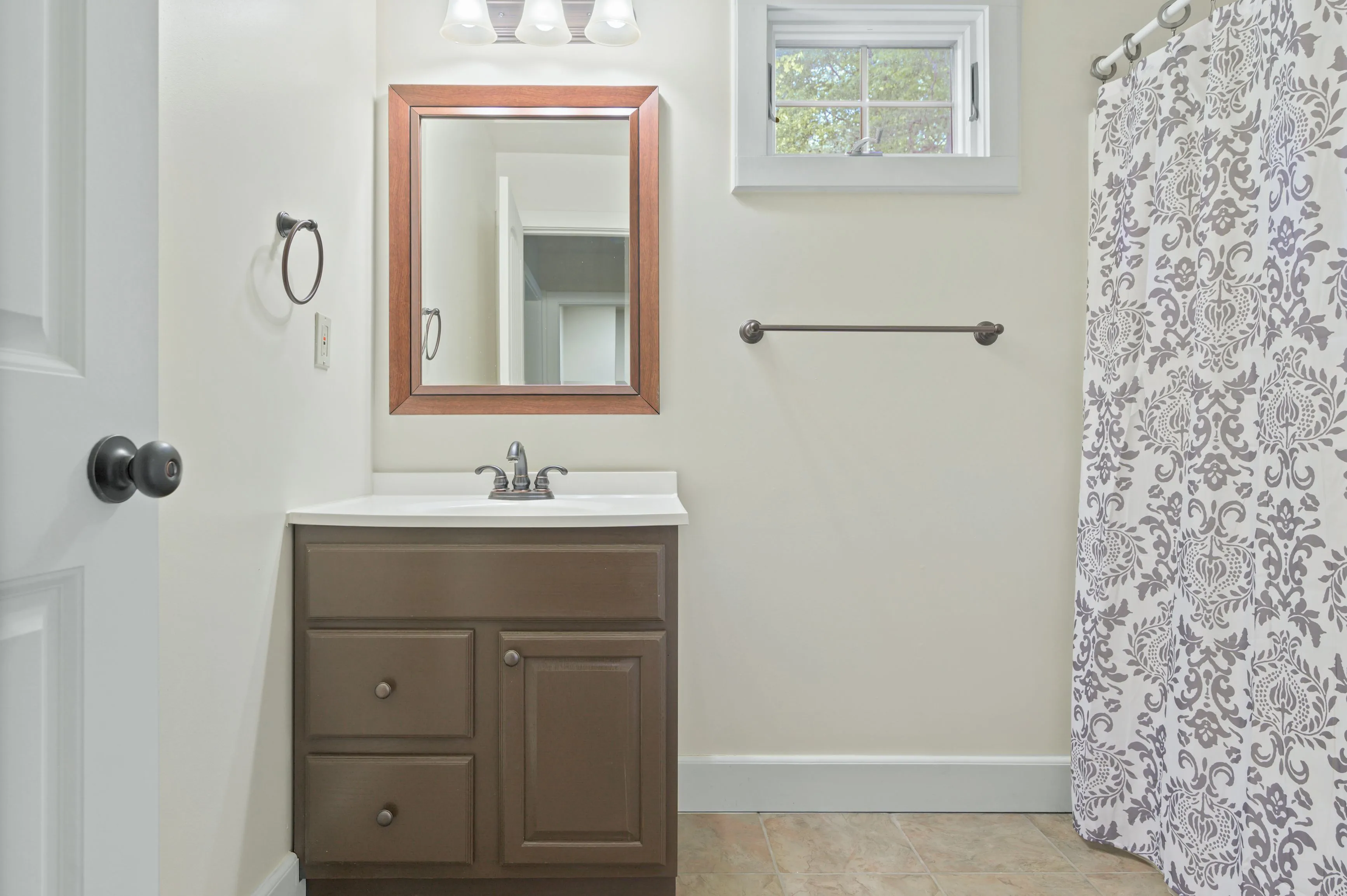 A modern bathroom with a white countertop, brown cabinets, a framed mirror, a patterned shower curtain, and a small window bringing in natural light.