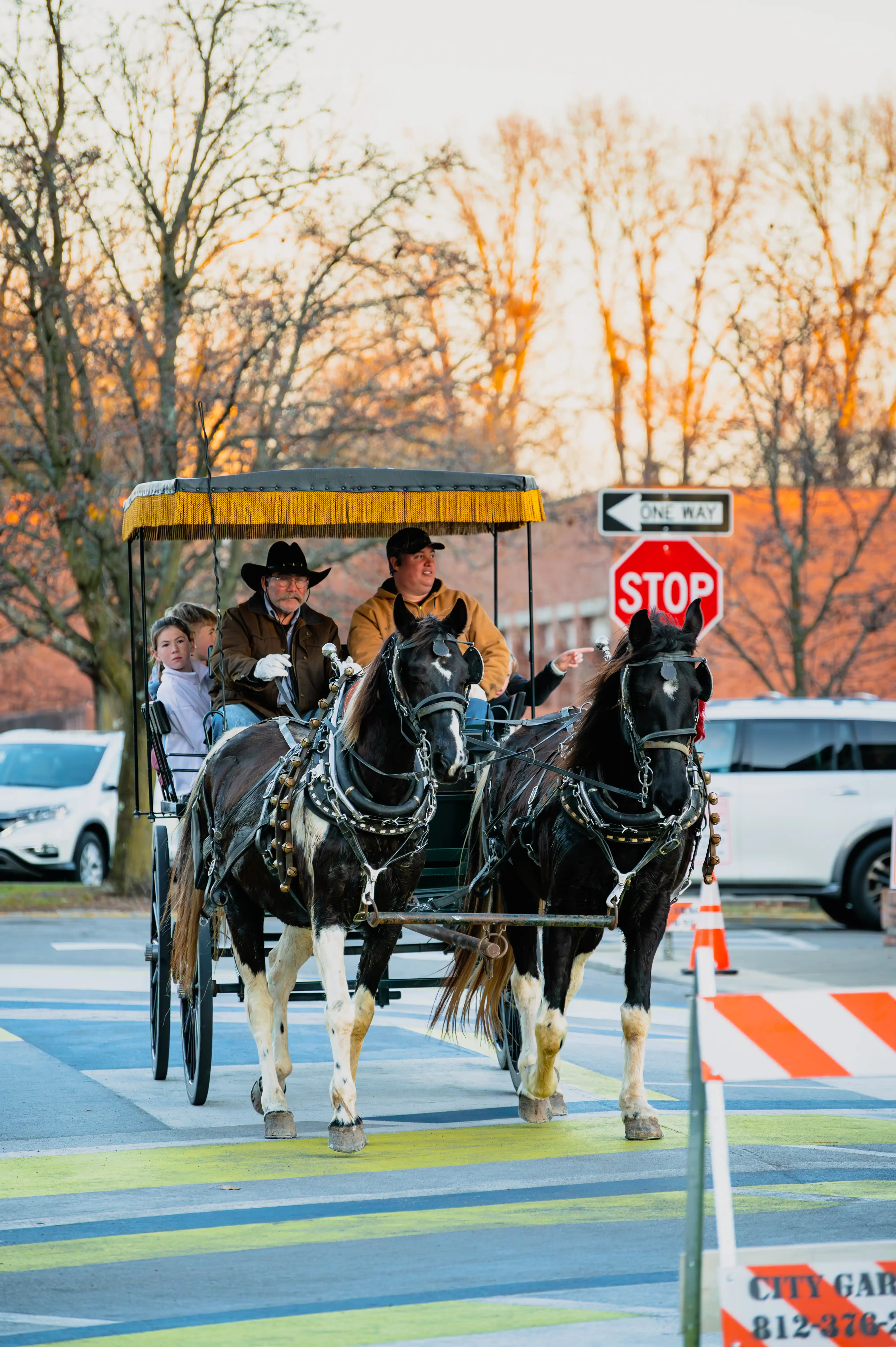 Horse-drawn carriage with passengers on a city street at dusk, crossing an intersection with a stop sign visible.