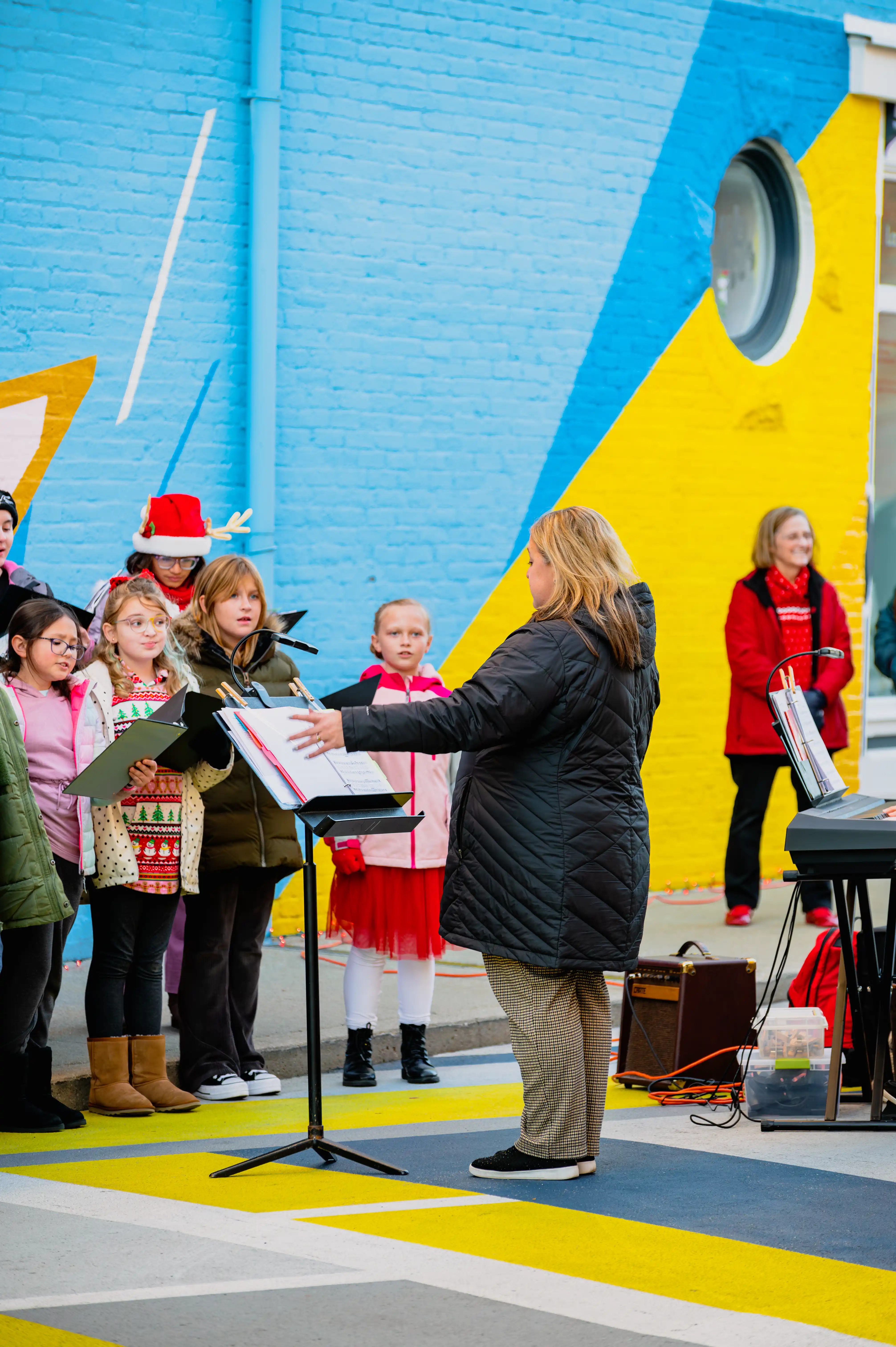Children's choir performing on stage with a conductor and keyboard player in front of a vibrant colorful backdrop.