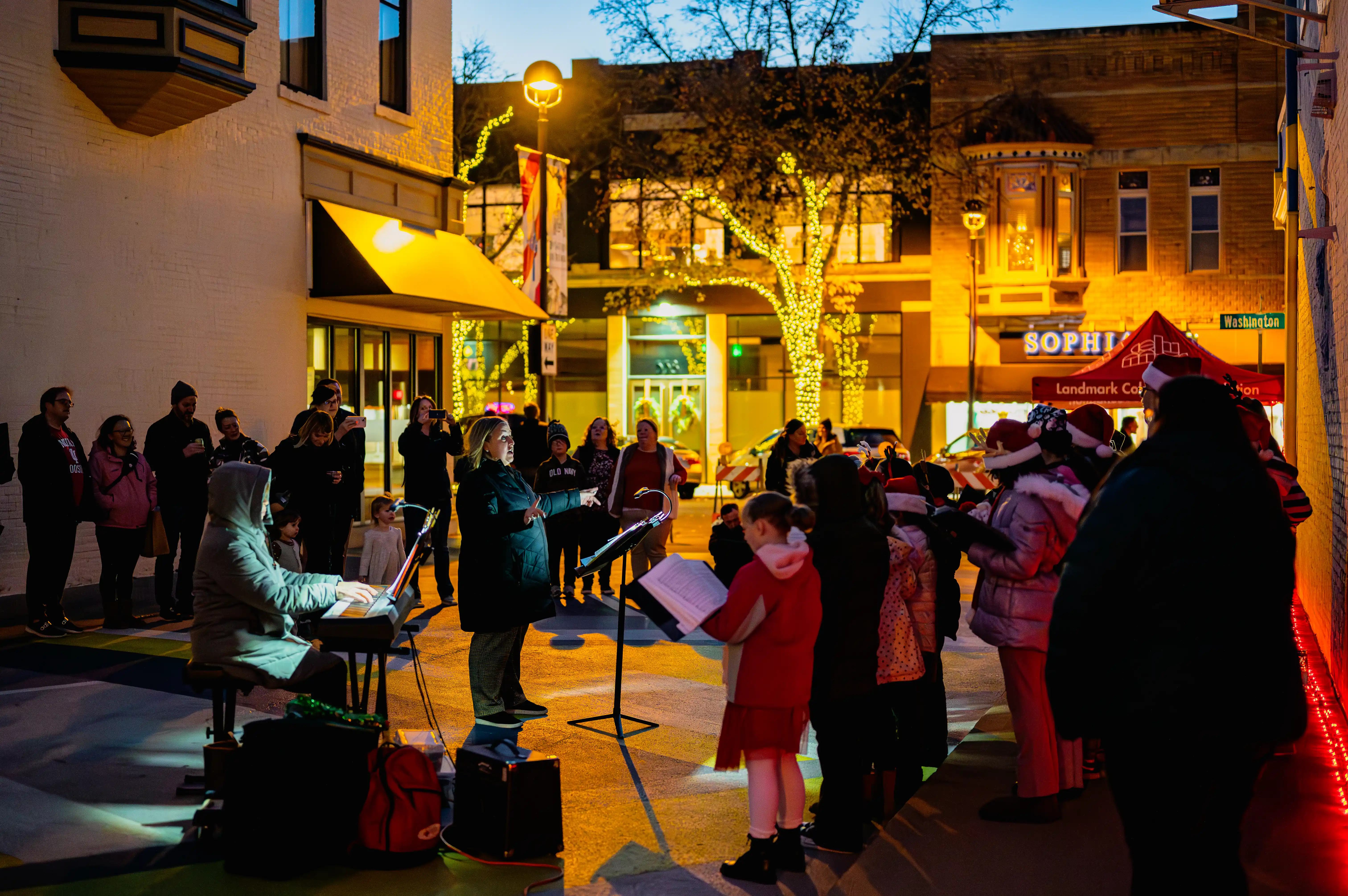A group of people gathered for an outdoor evening event with someone playing a keyboard, surrounded by festive lights and holiday decorations.