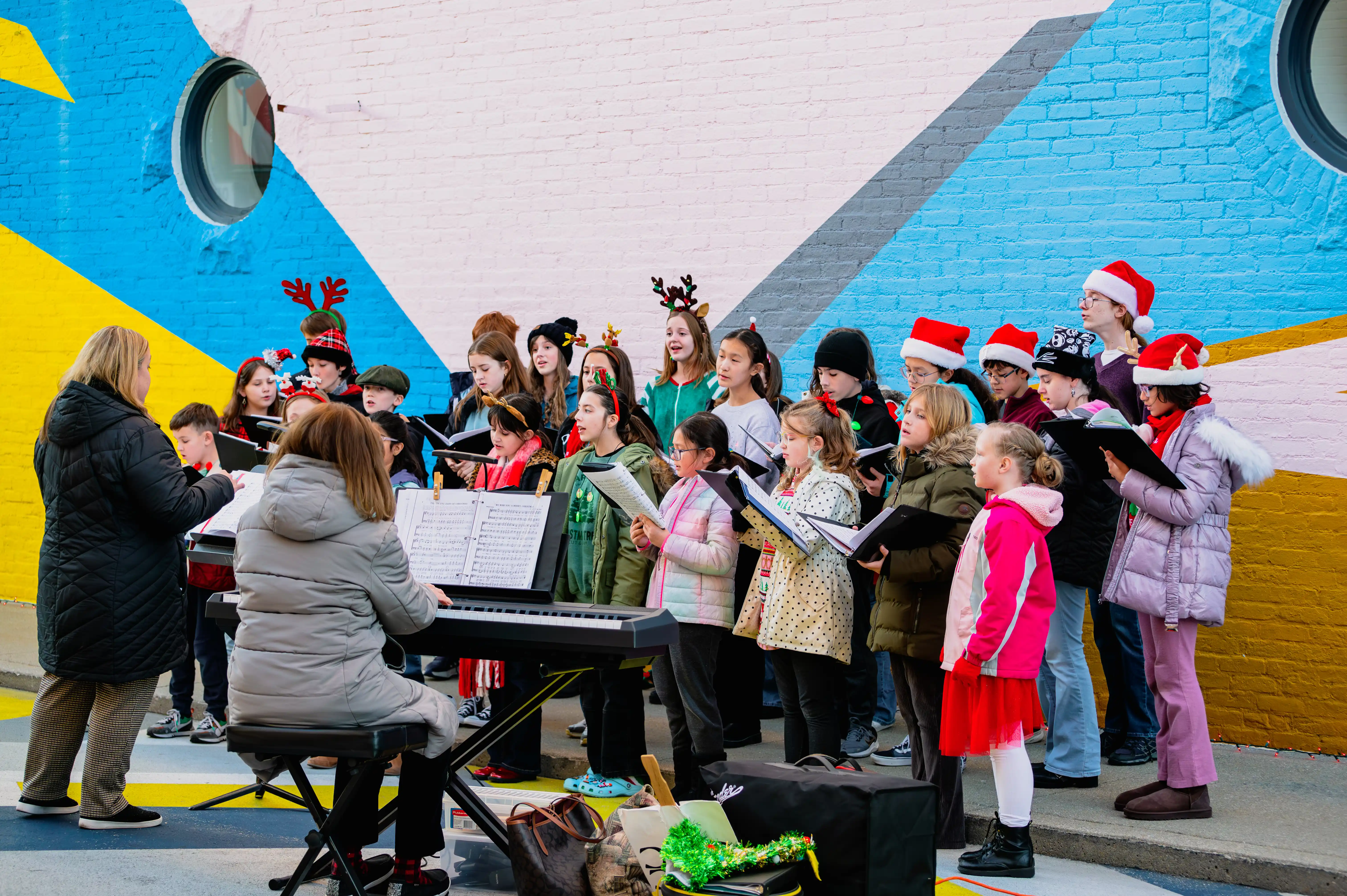 Children's choir performing outdoors with a conductor and pianist, wearing festive holiday attire including Santa hats and reindeer antlers.