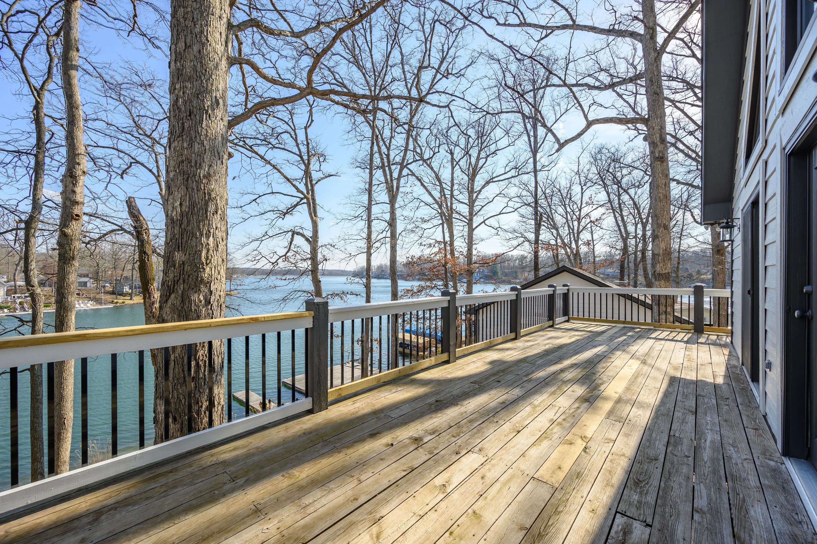 Sunny lakeside wooden deck with railing next to a house, surrounded by bare trees with a clear blue sky.