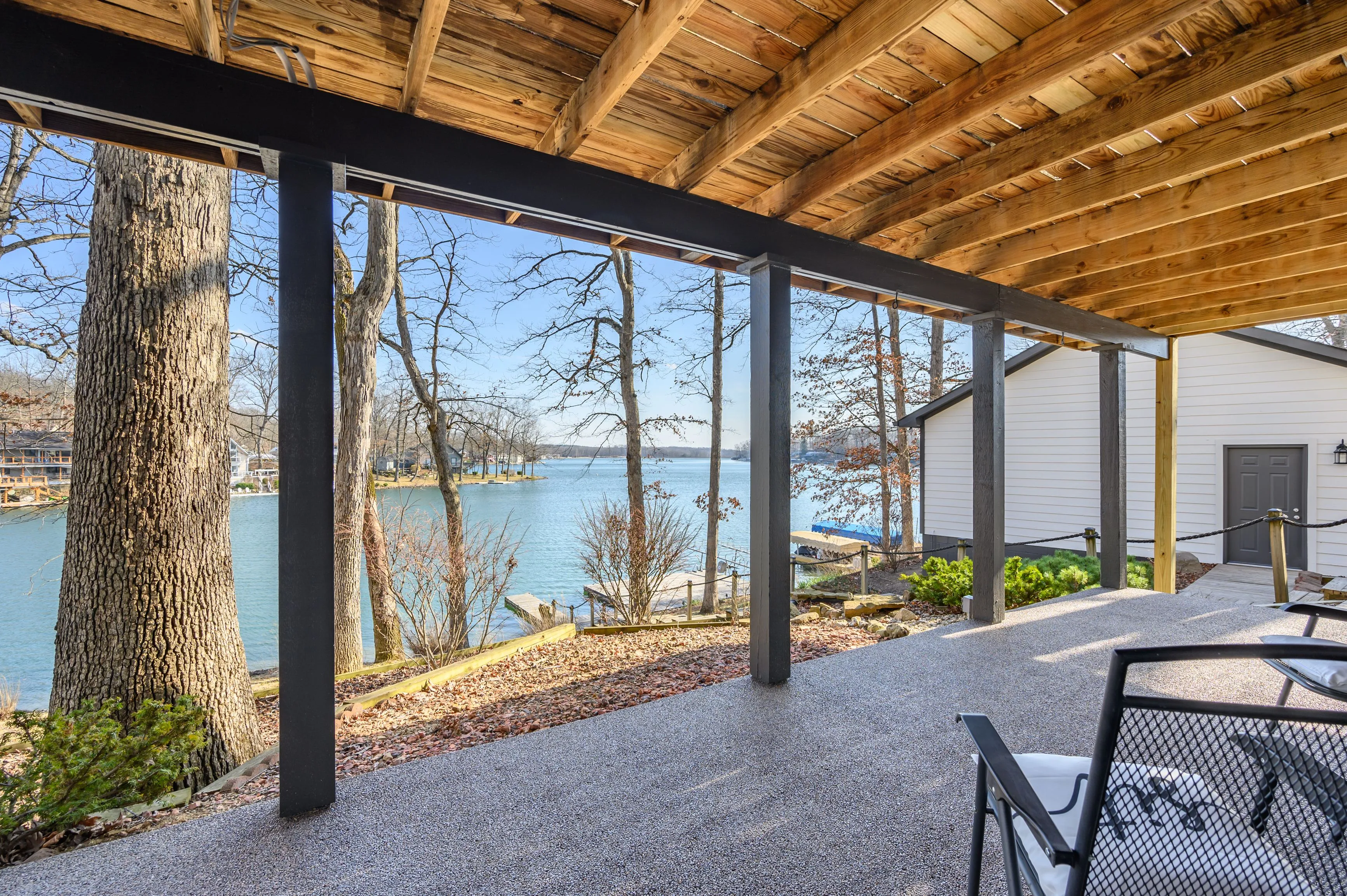 "View from a covered patio overlooking a tranquil lake with trees and clear skies, featuring outdoor furniture and wooden beams"