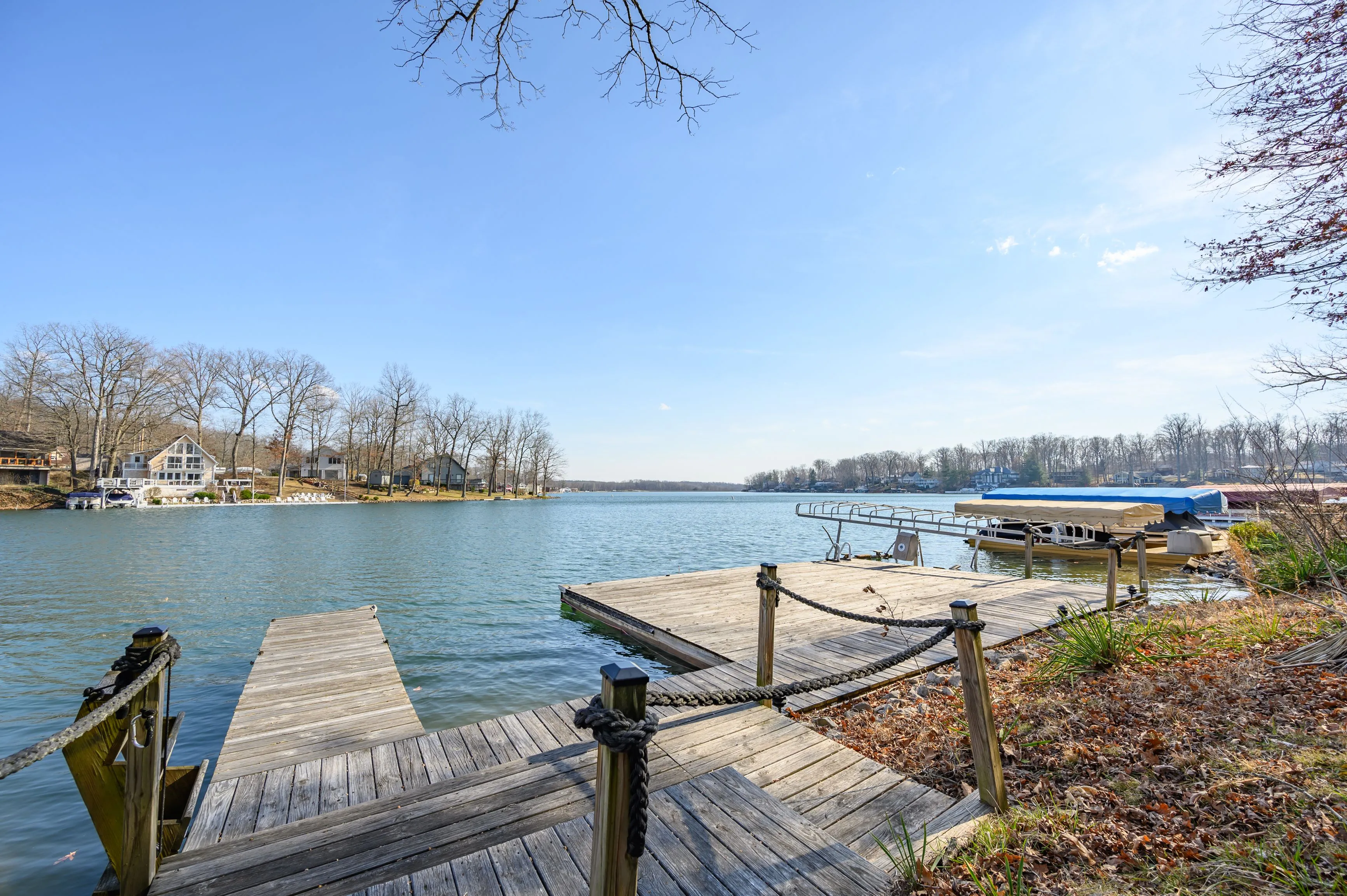 Wooden dock overlooking a tranquil lake with a boat cover on the right and residential houses in the background under a clear blue sky.