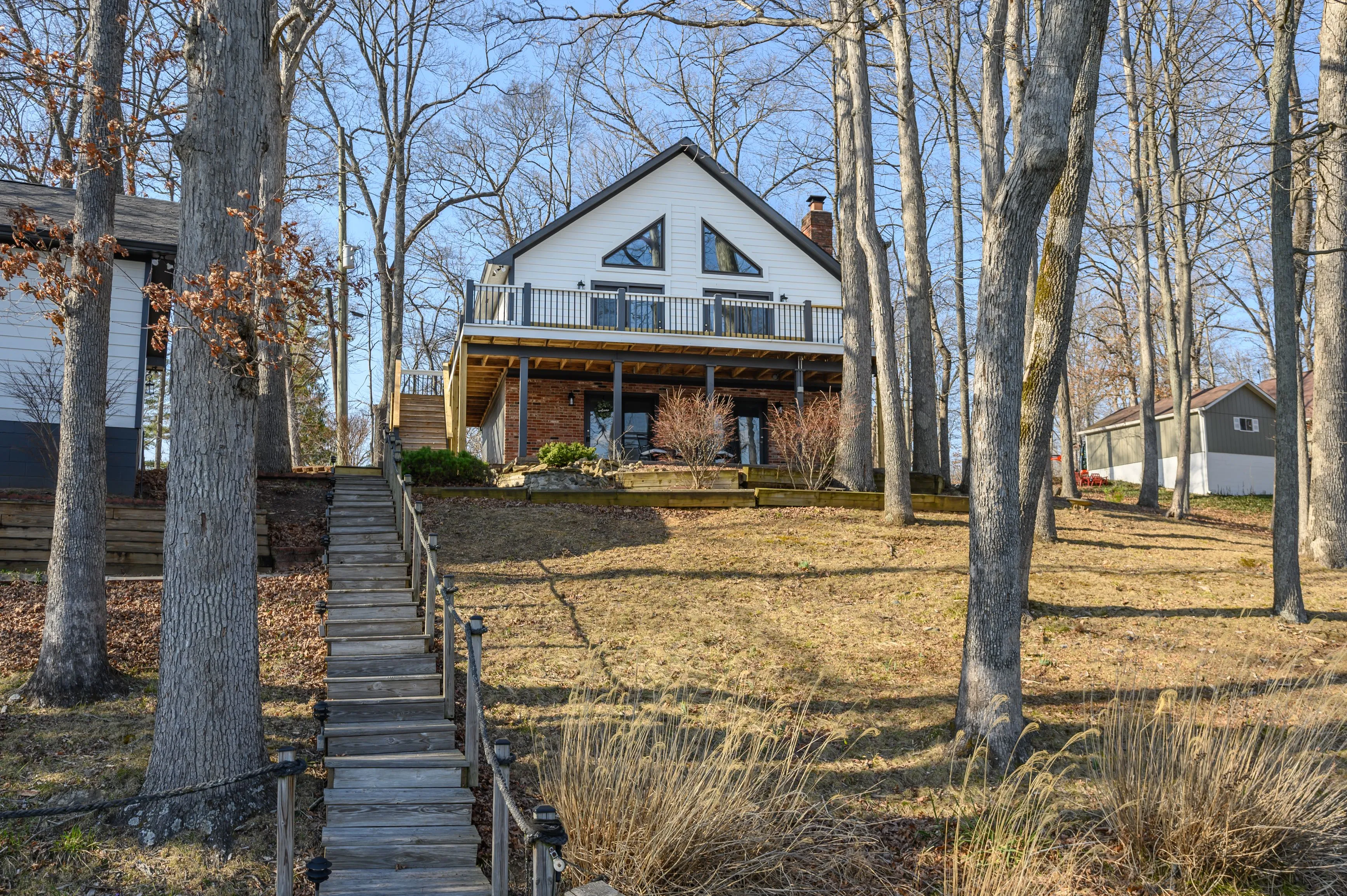 A two-story house with a brick foundation and white siding, featuring a large front porch, set back from a leafless tree-lined yard with a central staircase leading to the front door.