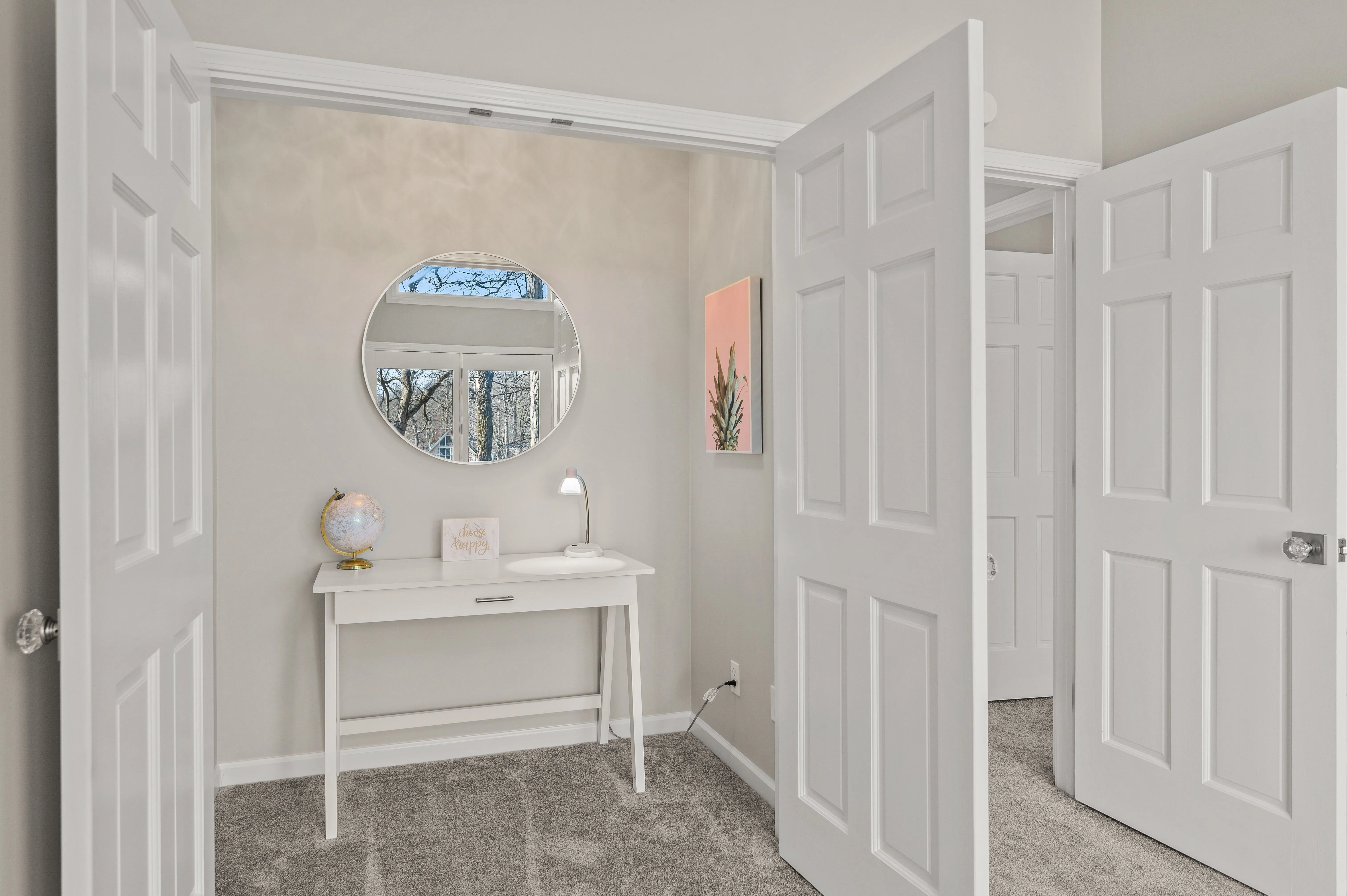 Brightly lit bathroom interior with an open door, featuring a white vanity with a round mirror, wall sconces, and decorative items on the countertop.
