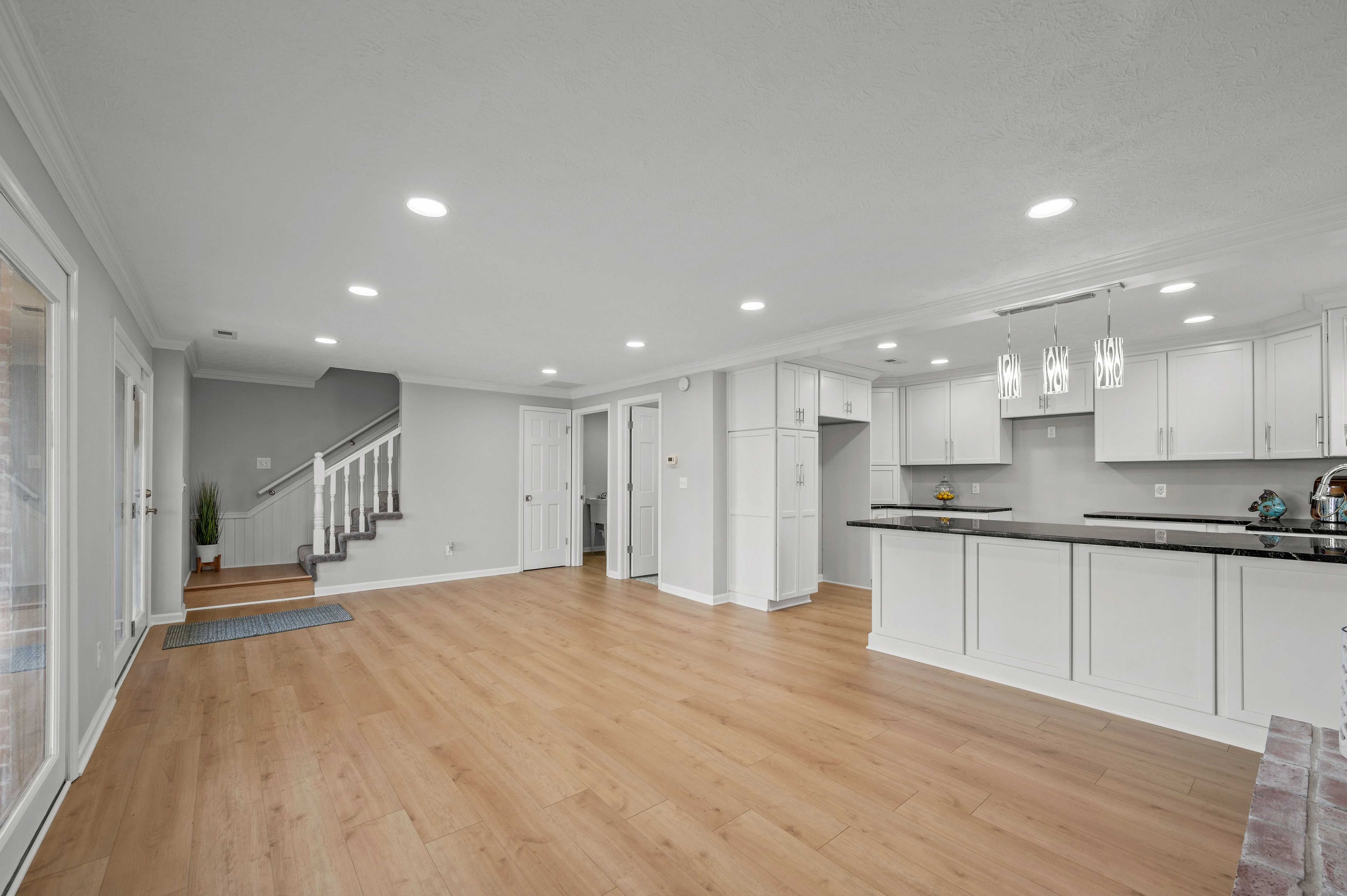 Spacious modern kitchen with white cabinets, stainless steel appliances, and hardwood floors leading to a living area with recessed lighting and a staircase on the left.