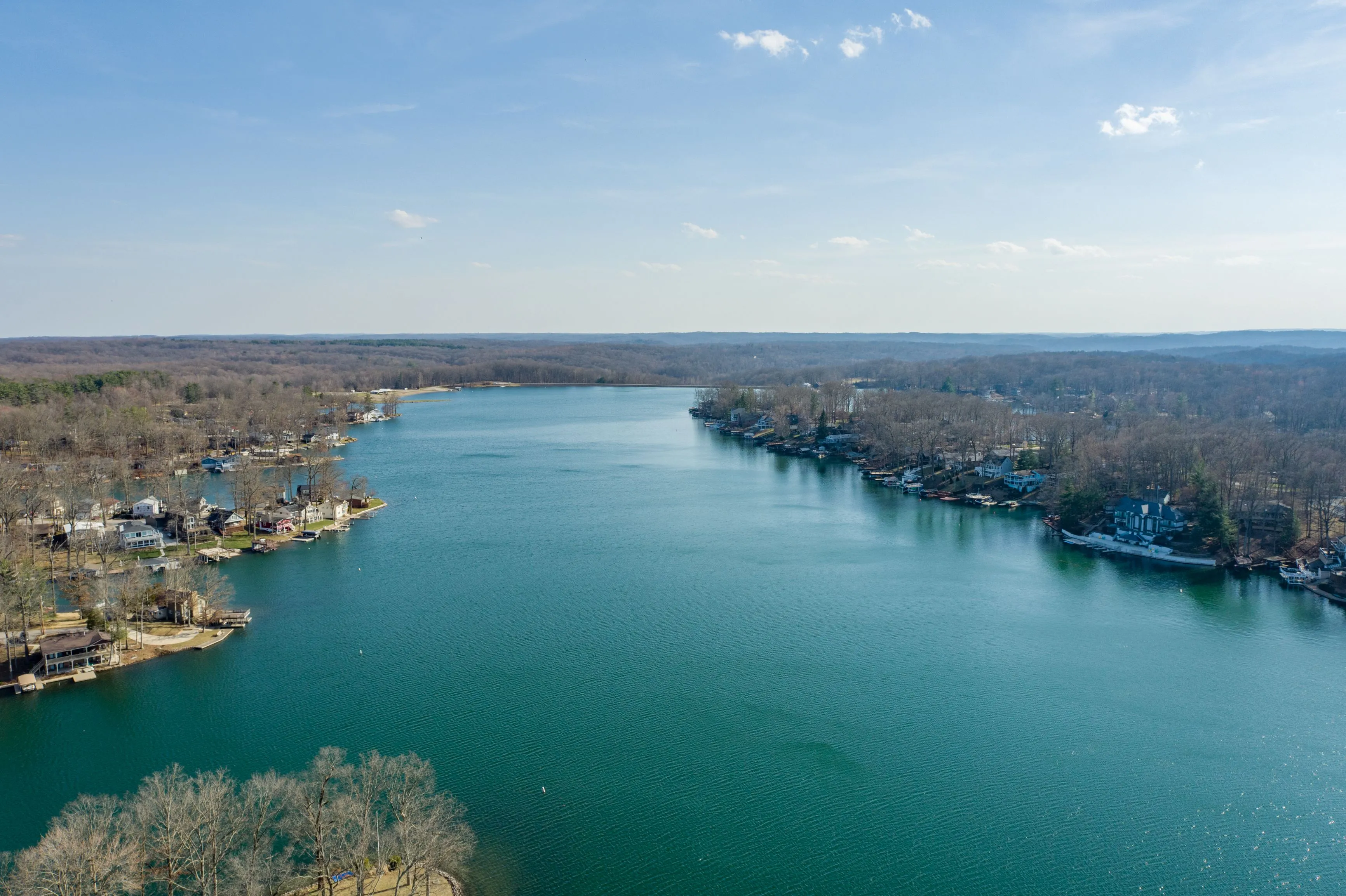 Aerial view of a serene lake with surrounding residential areas and dense trees under a clear blue sky.