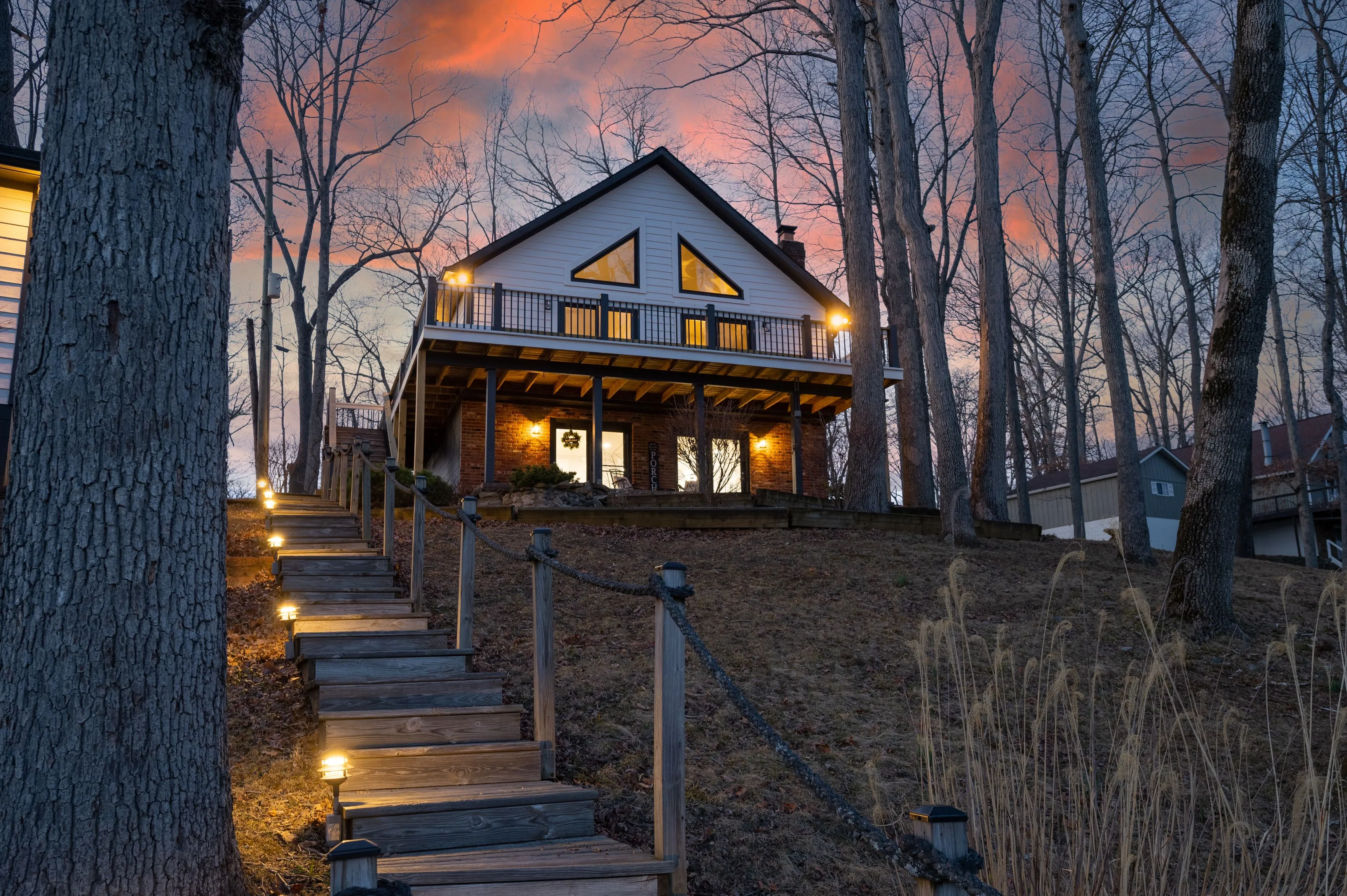 Twilight scene featuring a cozy two-story house with lit windows at the end of a stairway flanked by leafless trees under a pink-hued sky.
