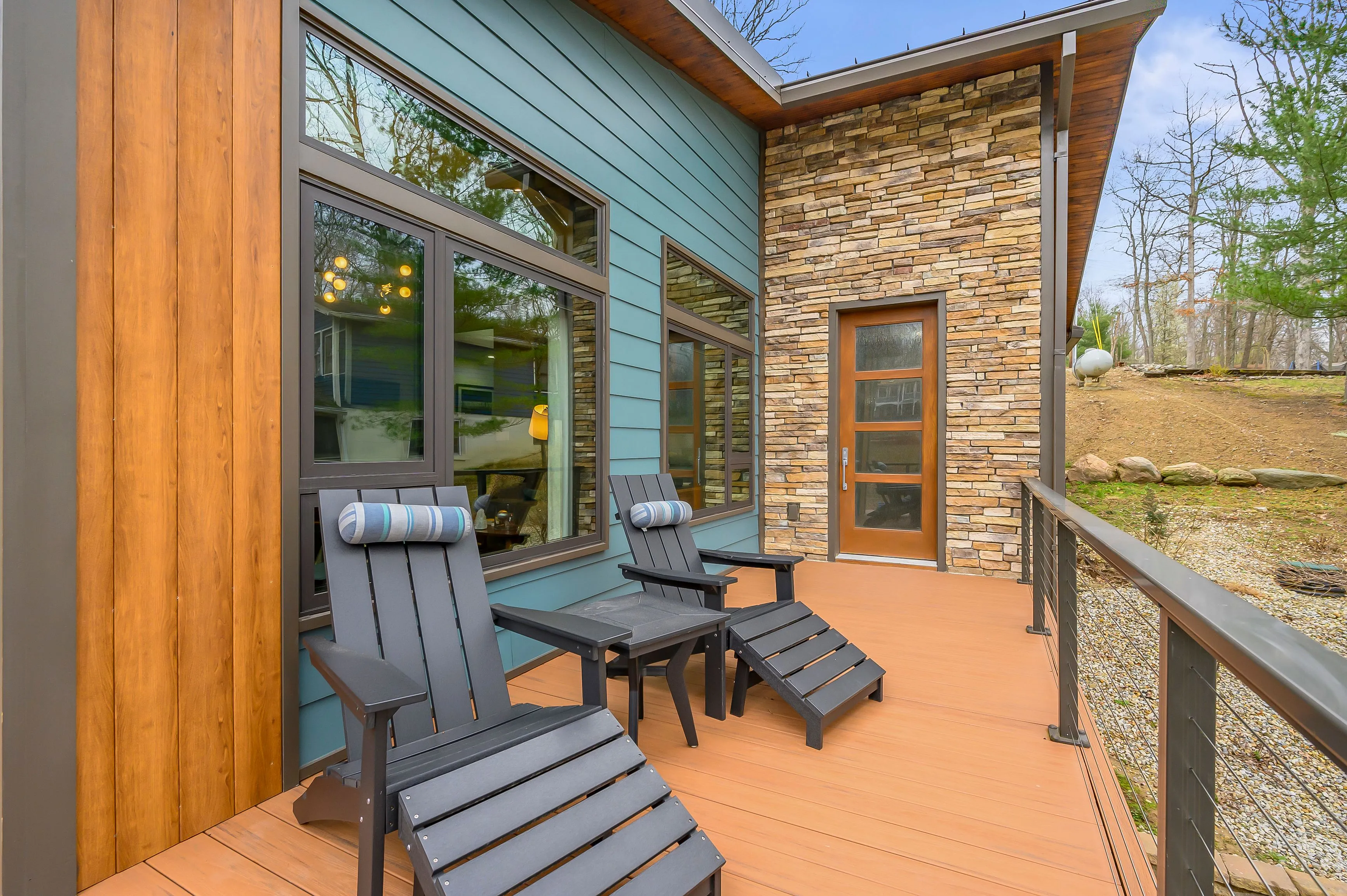 Cozy patio area with two Adirondack chairs and a small table on a wooden deck, adjacent to a house with blue siding and stone accents.