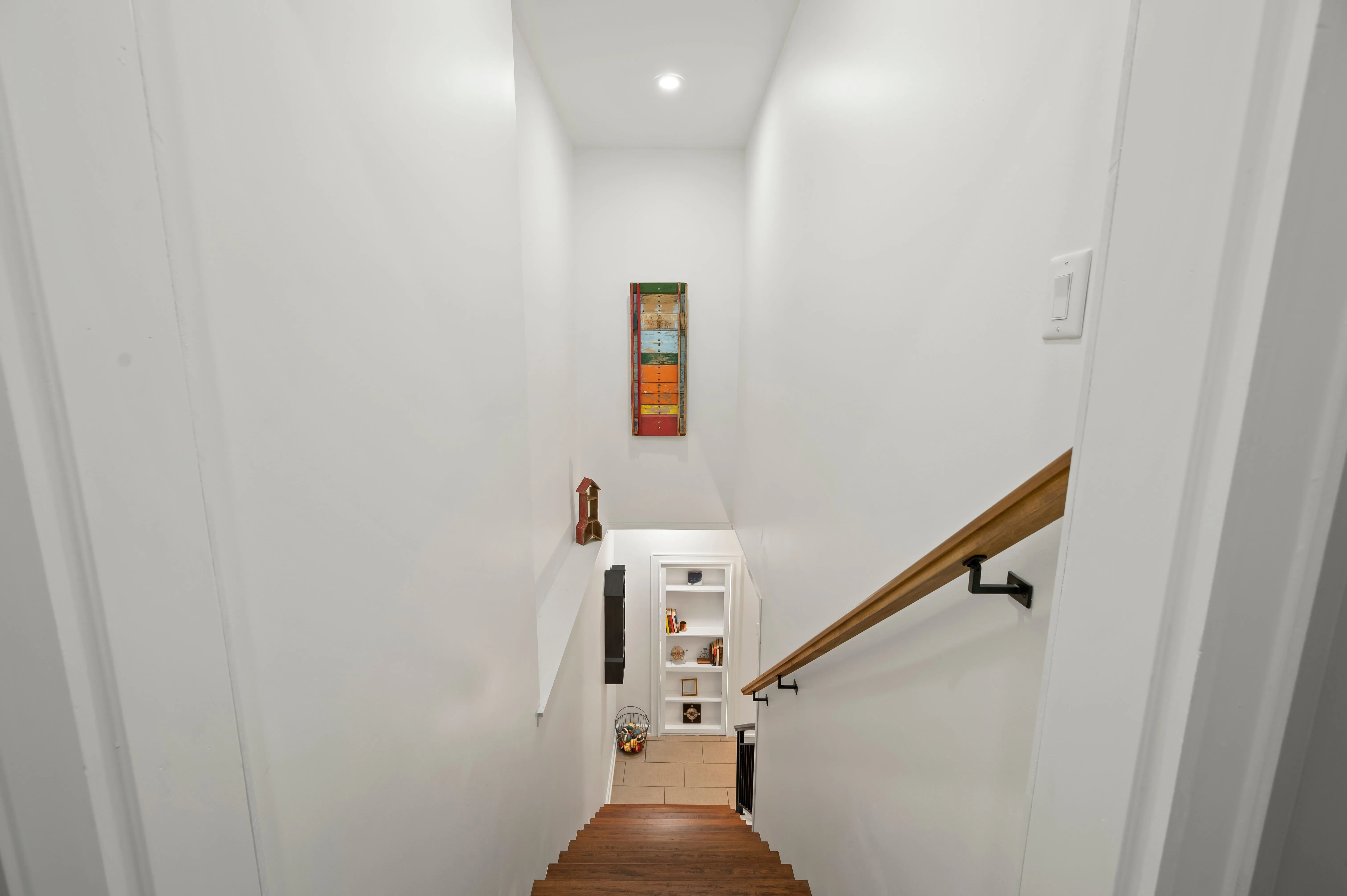 Narrow staircase with white walls, wooden steps, and a handrail leading down to a door.