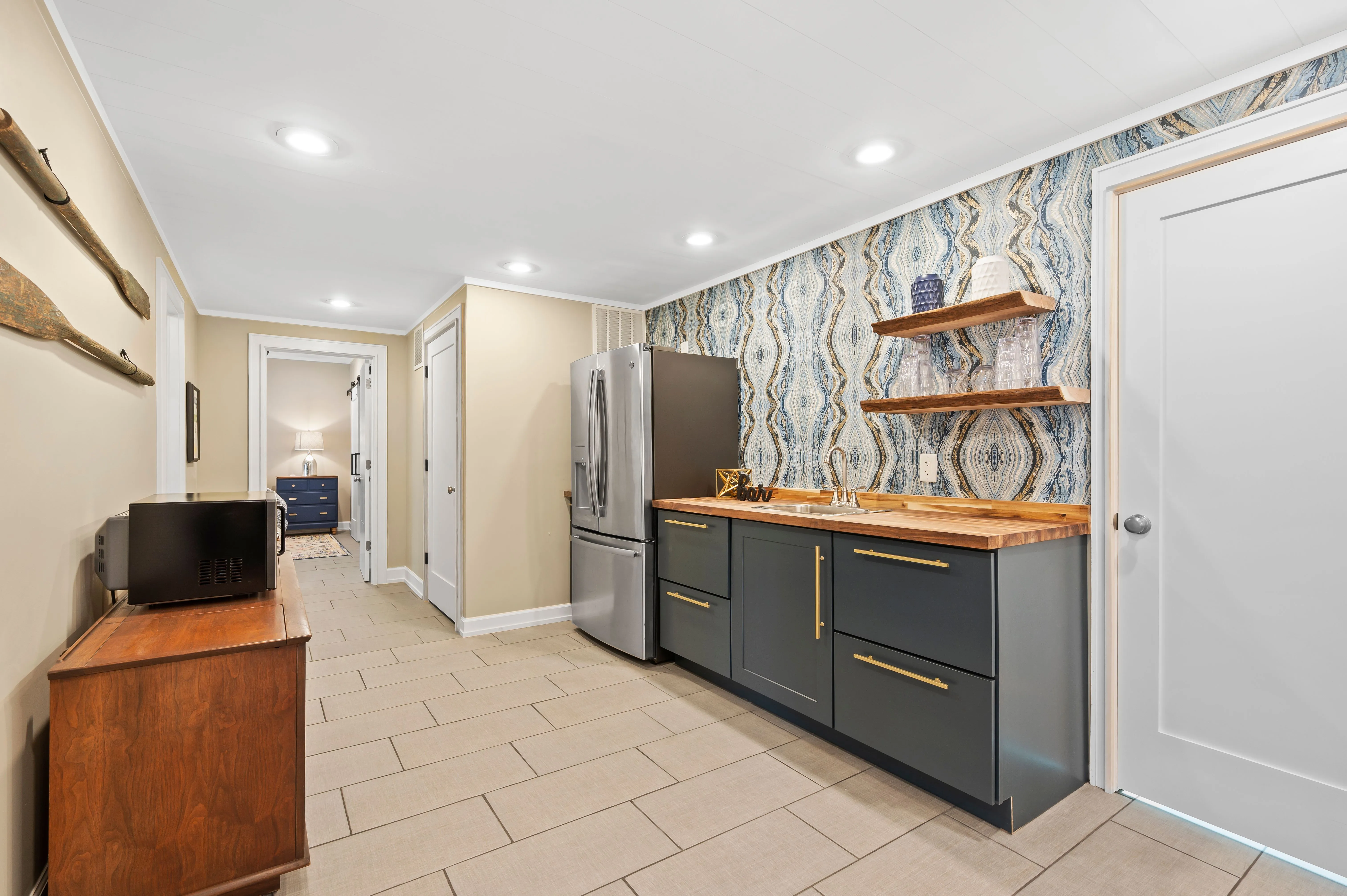 Modern hallway interior with decorative blue pattern wallpaper, dark kitchen cabinetry, wooden shelves, and tiled floor.