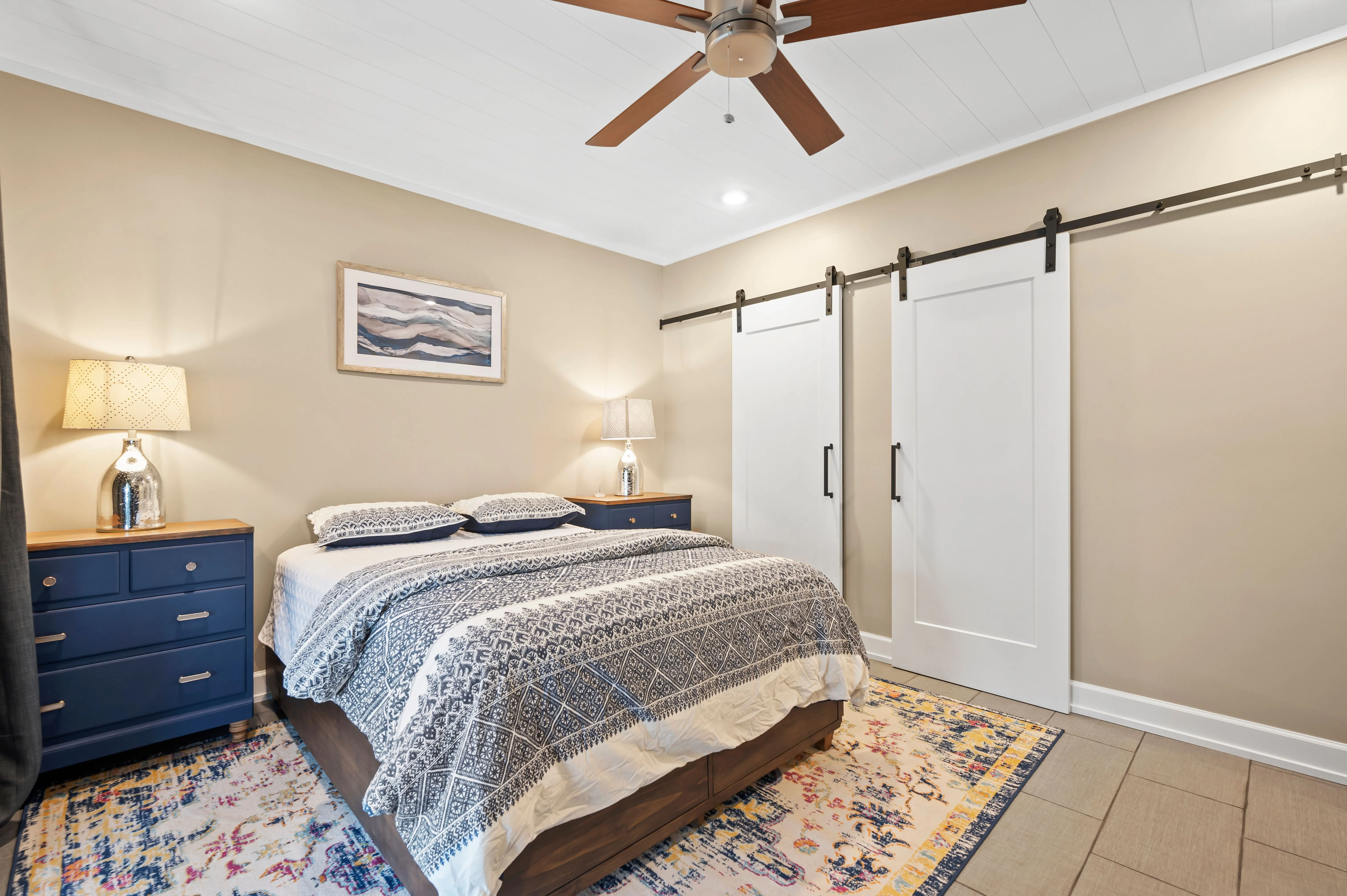 Cozy bedroom interior with a queen-size bed, patterned bedding, a ceiling fan, sliding closet doors, and a nightstand with a lamp.