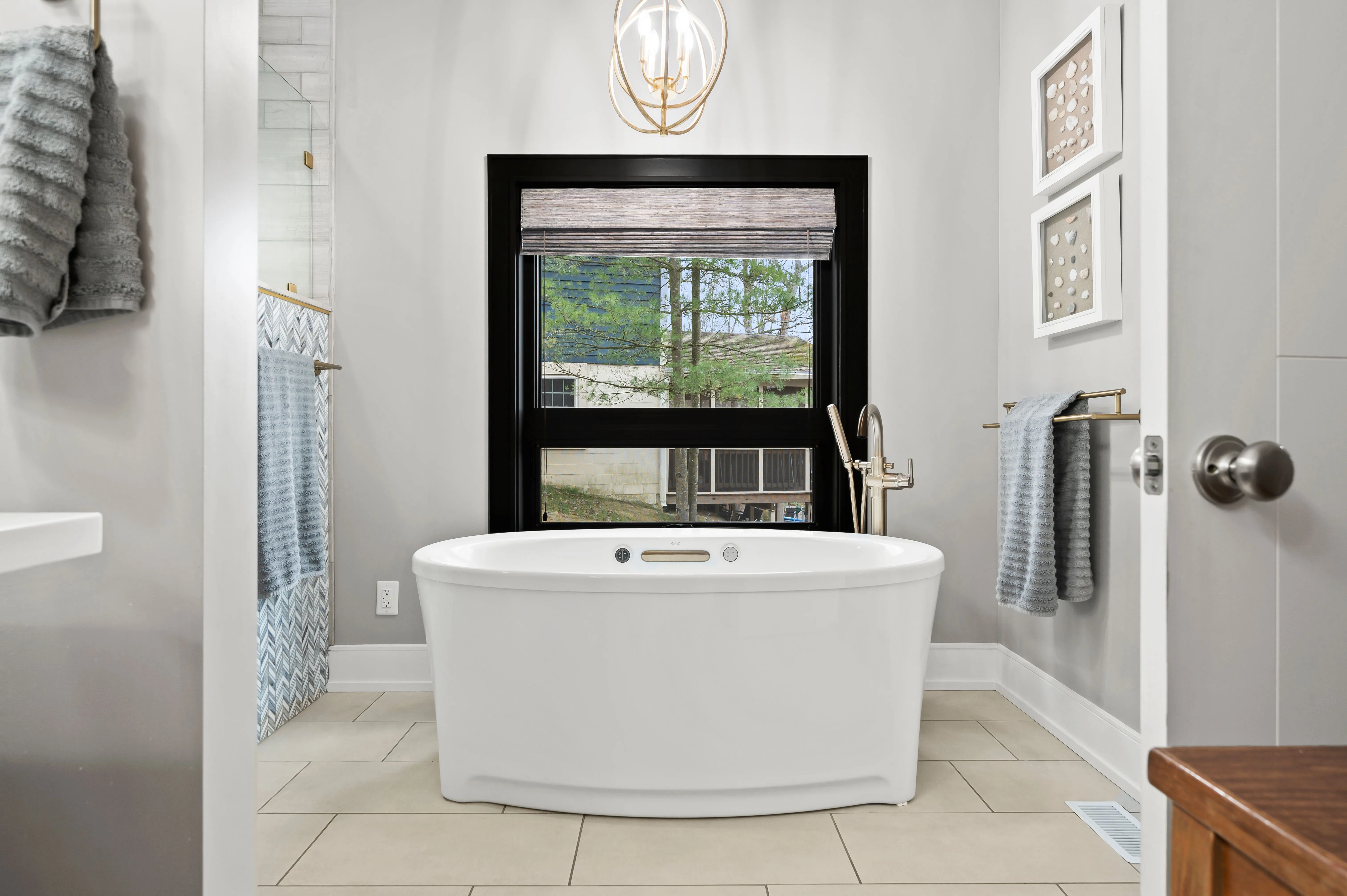 Modern bathroom interior with a freestanding tub, gray tiles, black framed window, and elegant lighting fixtures.