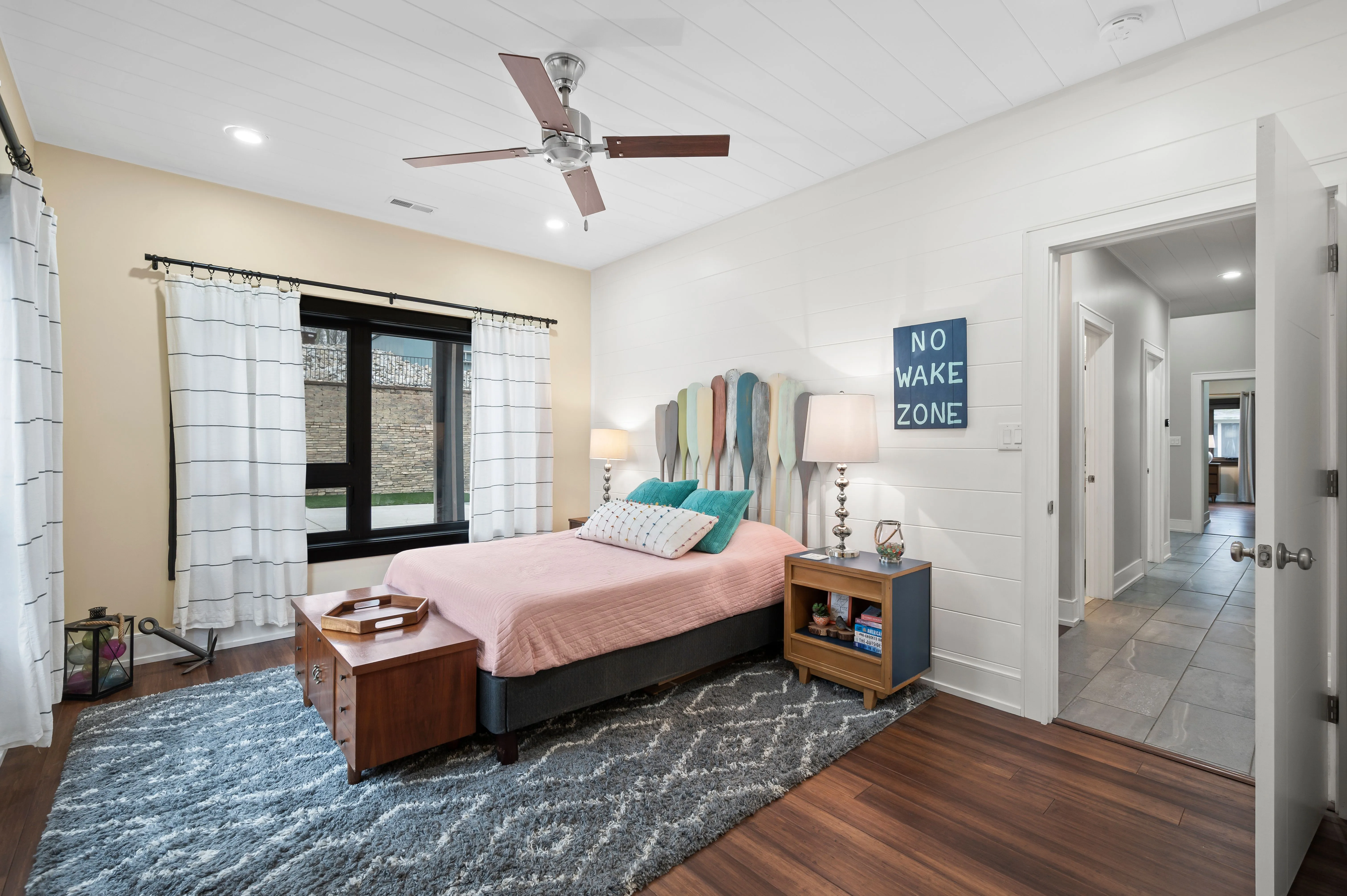 Bright and modern bedroom interior with a queen-sized bed, ceiling fan, hardwood floors, and a decorative area rug. The room features a large window with curtains, bedside tables with lamps, and wall art. A hallway entrance is visible on the right.