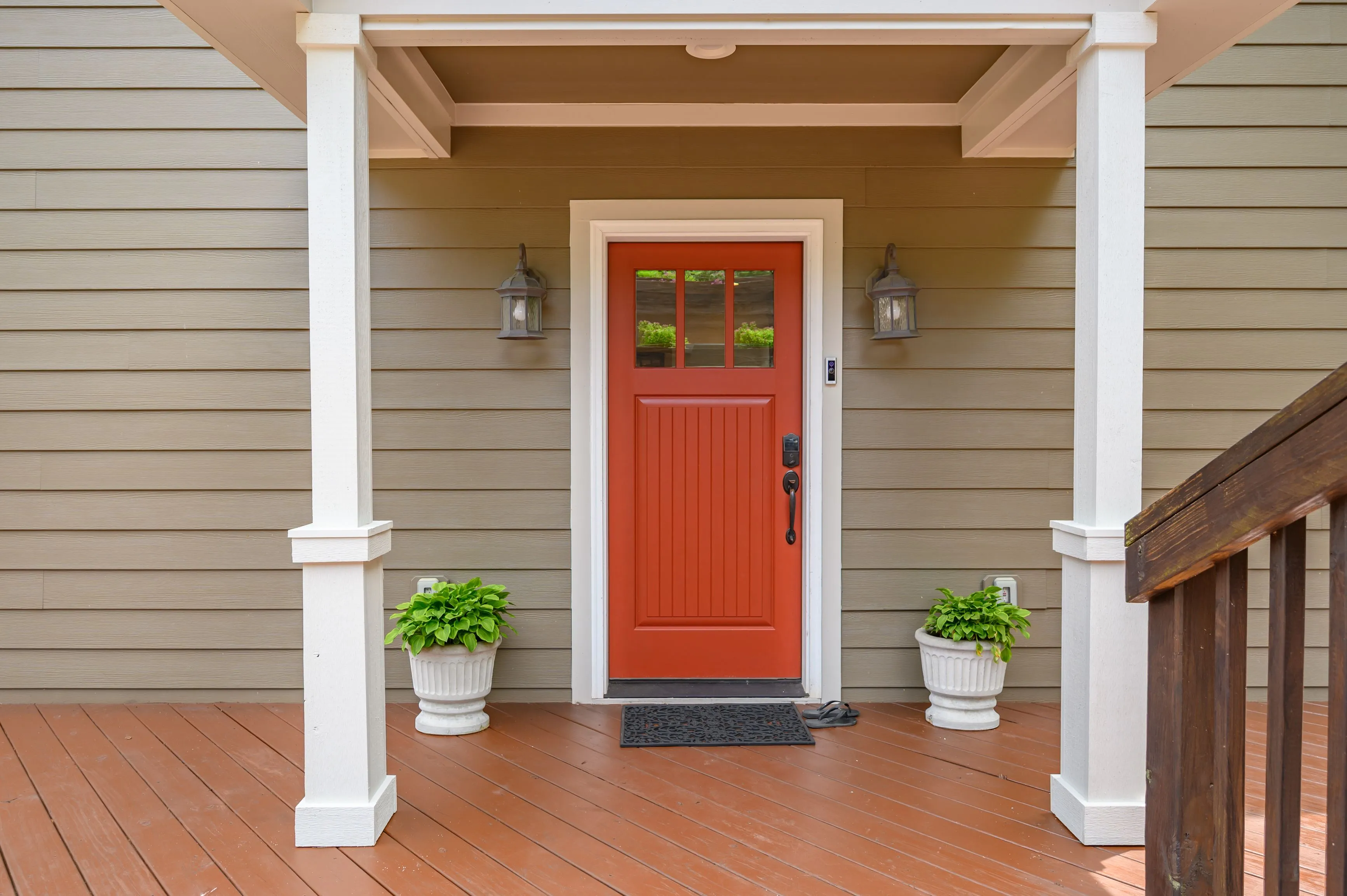 Front porch of a house with a red door, white columns, potted plants, and wooden decking.