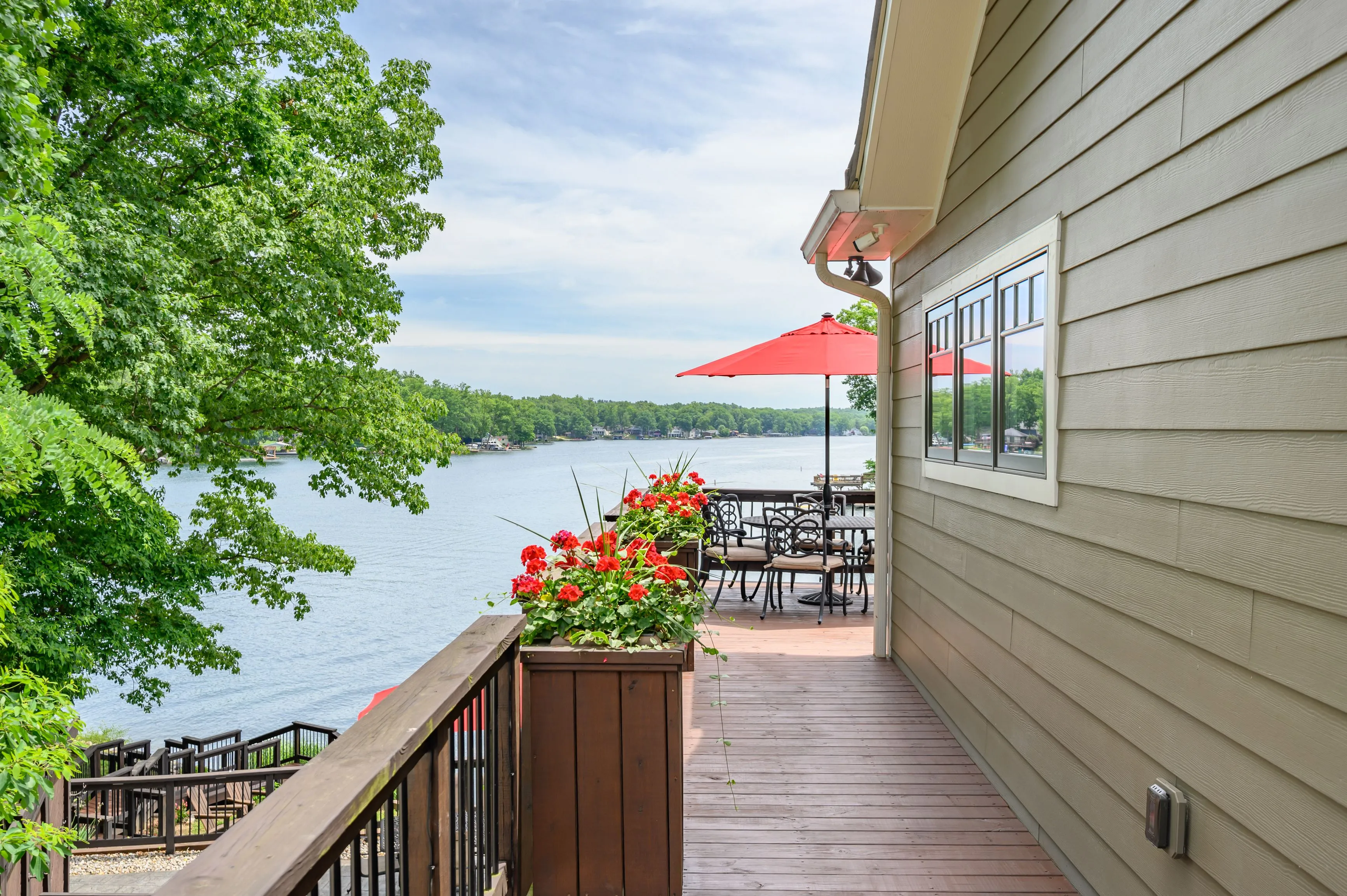 A scenic view from a lakeside home's deck featuring a dining area under a red umbrella, surrounded by lush greenery, overlooking a tranquil lake.