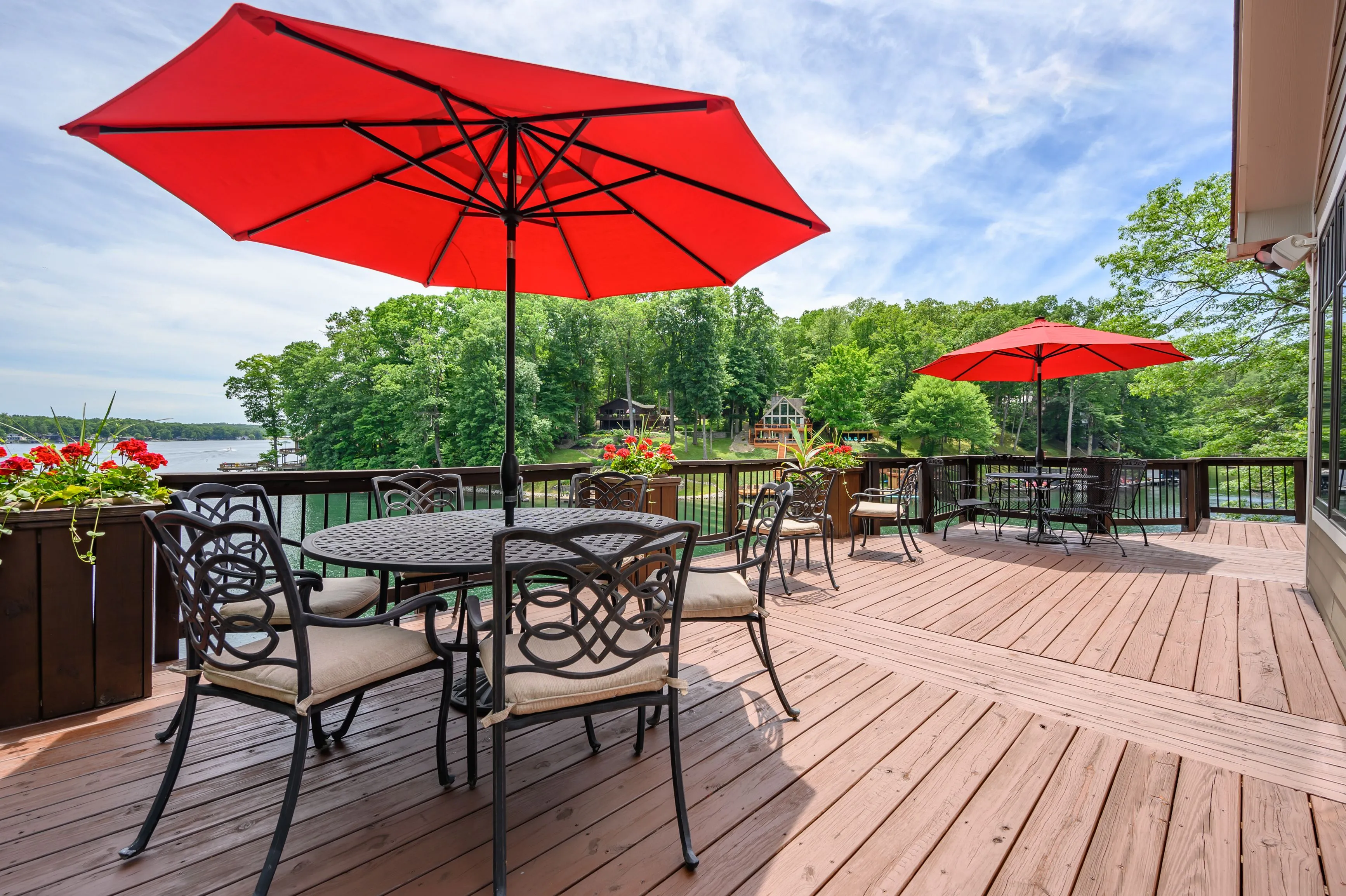 A lakeside deck with outdoor dining furniture under red umbrellas on a sunny day.