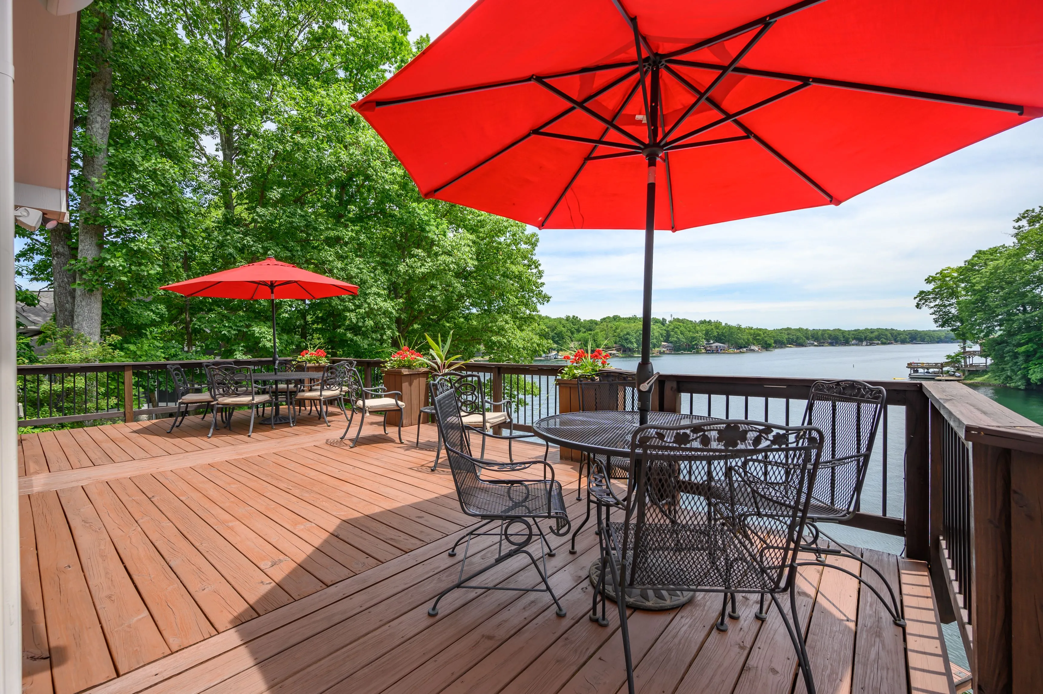 Lakeside wooden deck with outdoor furniture and a red umbrella, surrounded by lush greenery.