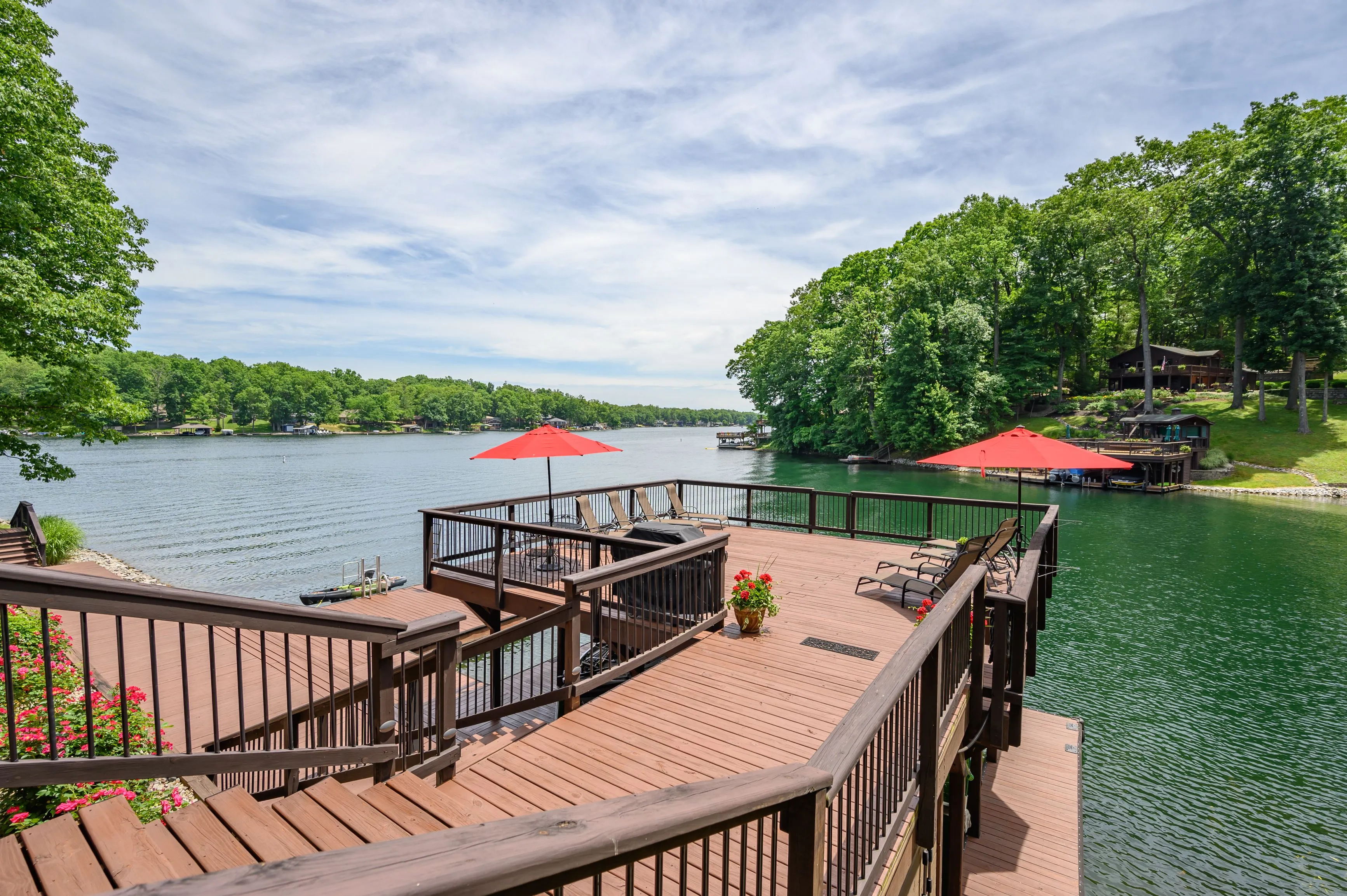 "Wooden deck with patio furniture overlooking a tranquil lake with red umbrellas and a lush green treeline under a clear sky"