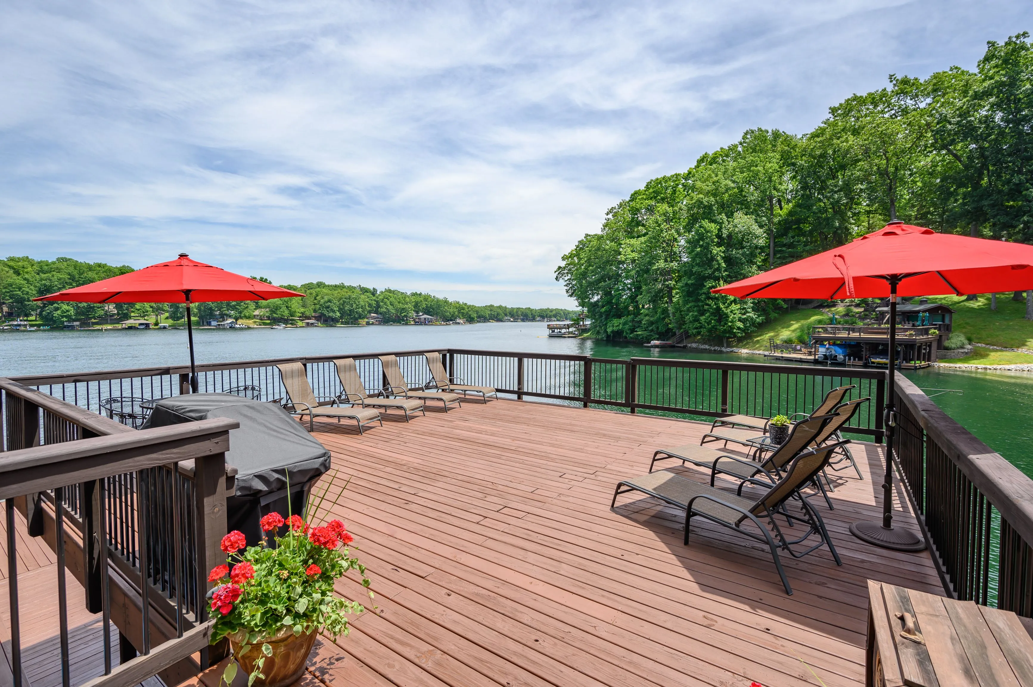 Wooden deck overlooking a tranquil lake with two red patio umbrellas, lounge chairs, and a flowerpot with red flowers.