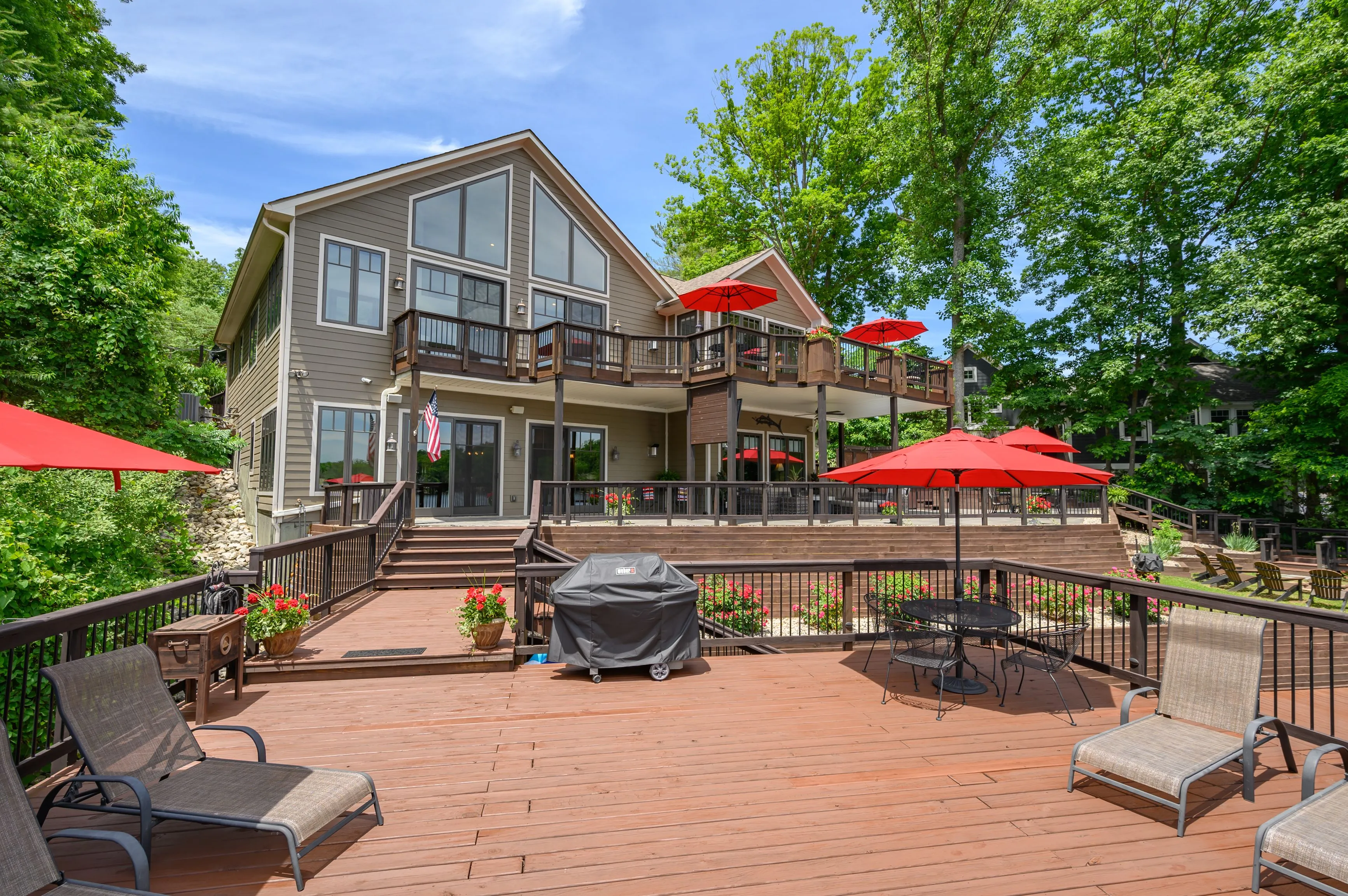 Spacious backyard patio with decking and outdoor furniture, featuring a barbecue grill and red umbrellas, with a two-story house surrounded by greenery in the background.