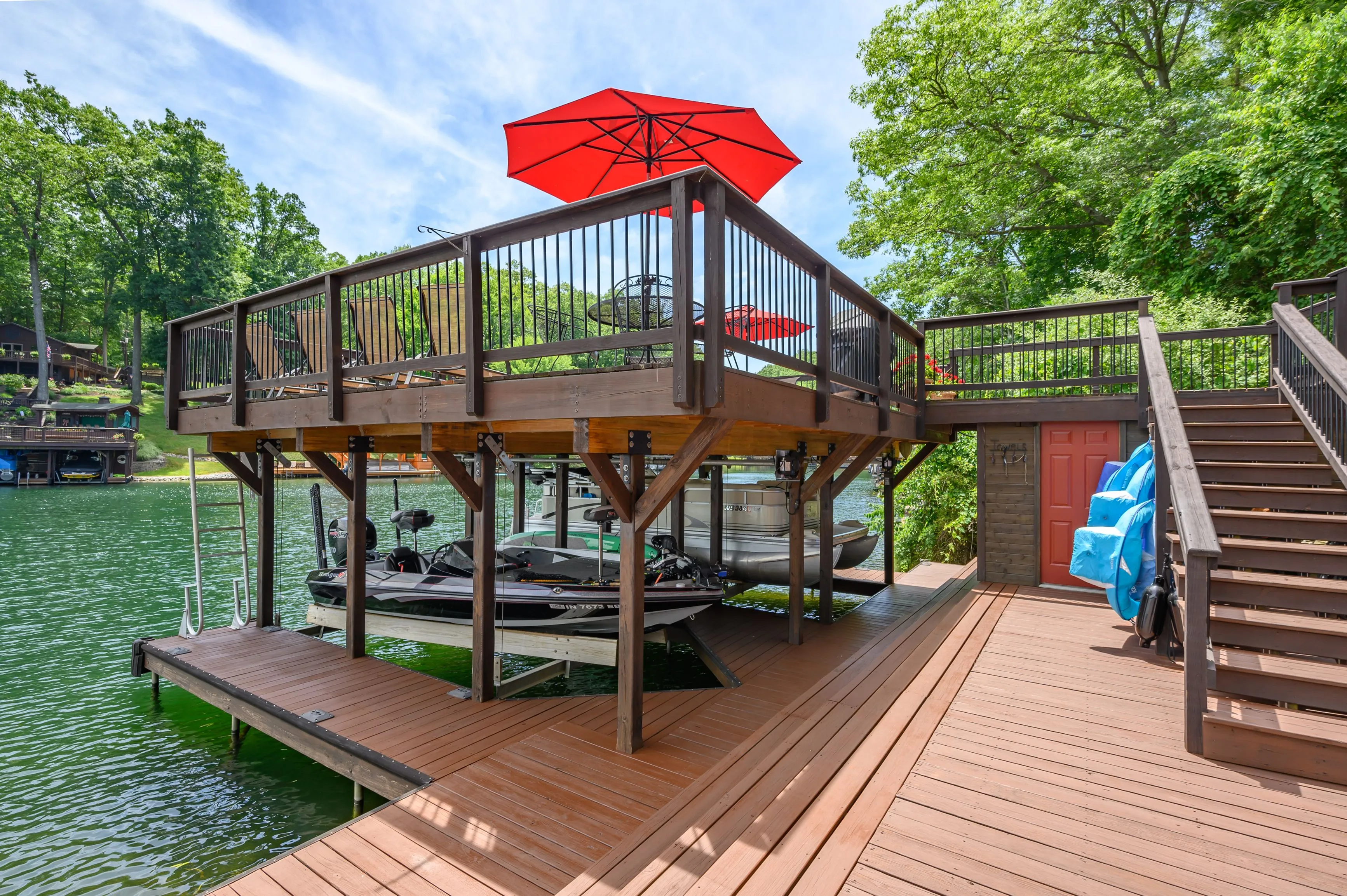 Lakeside wooden deck with a boat dock underneath an elevated patio area with metal railings and a red umbrella on a sunny day.
