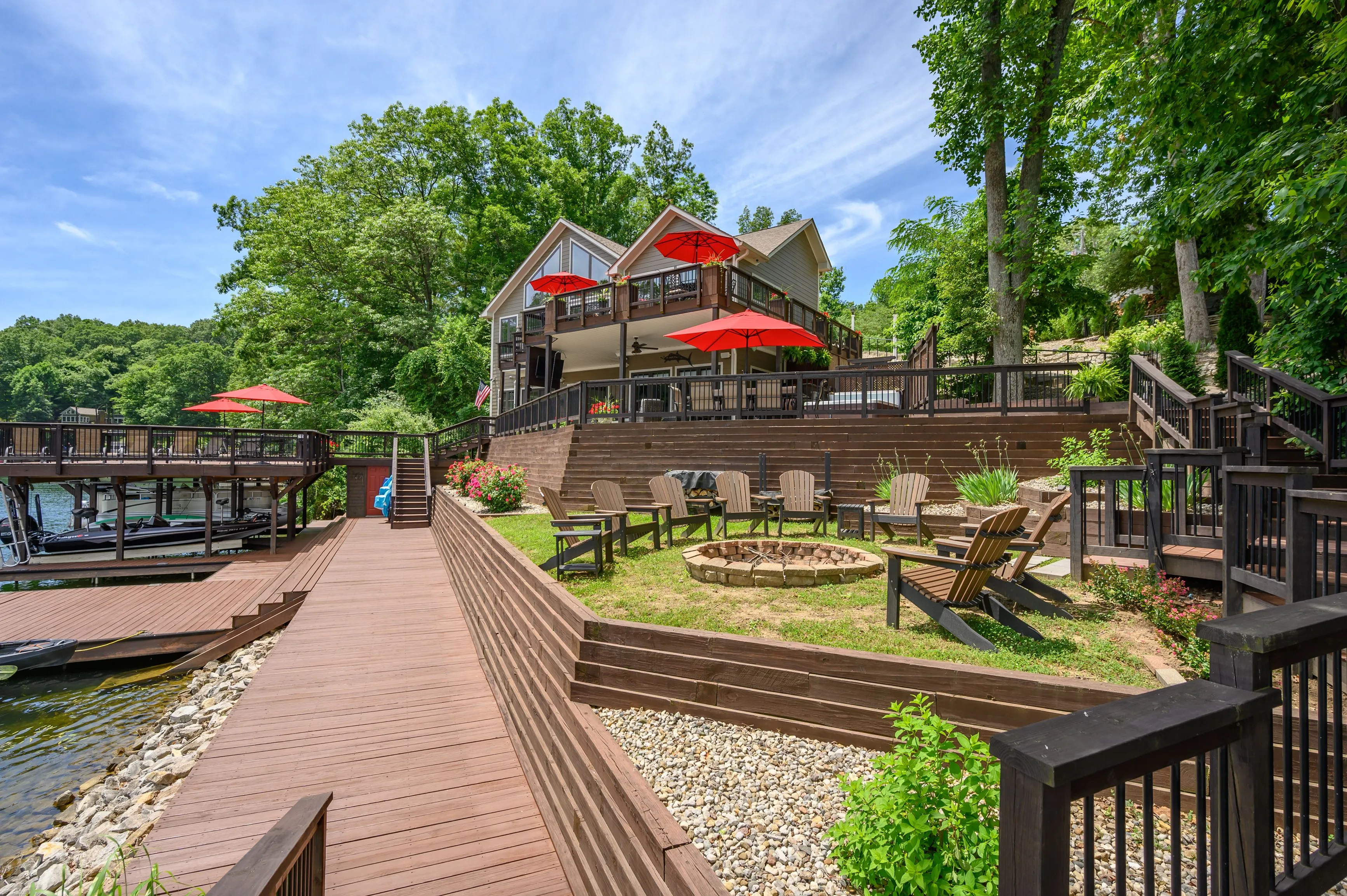 Lakeside house with multiple decks and red umbrellas surrounded by trees with a wooden dock leading up to the property.