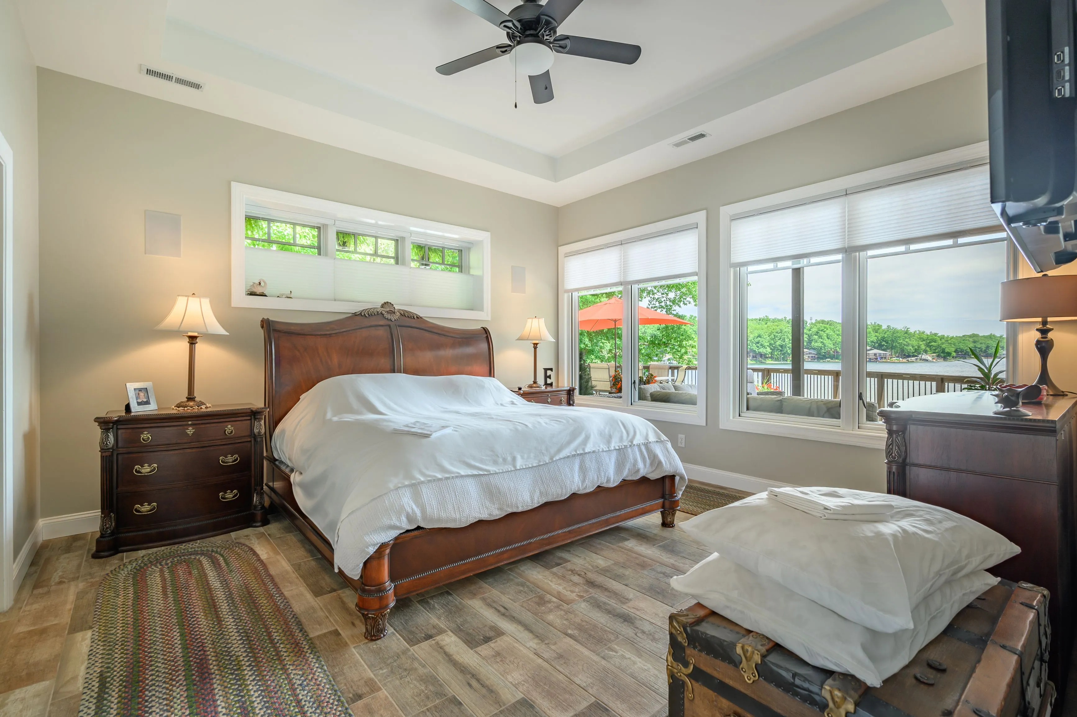 Bright and airy bedroom with a queen-sized bed, wooden furniture, ceiling fan, and large windows showing greenery outside.