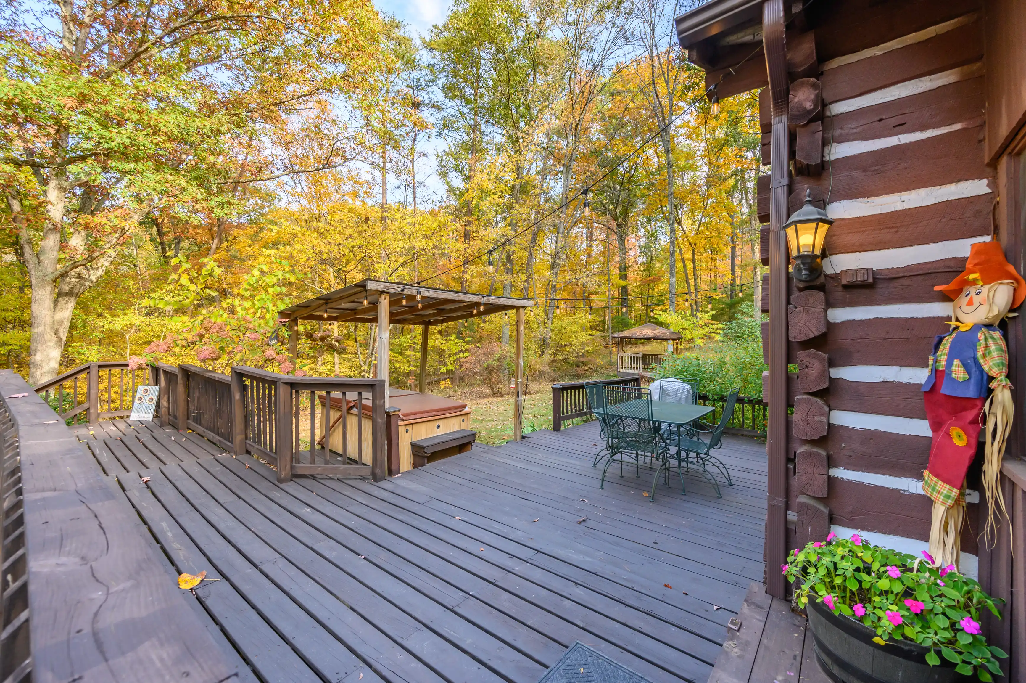Wooden deck of a rustic cabin with patio furniture, a gazebo, and decorative scarecrow, surrounded by autumn foliage.