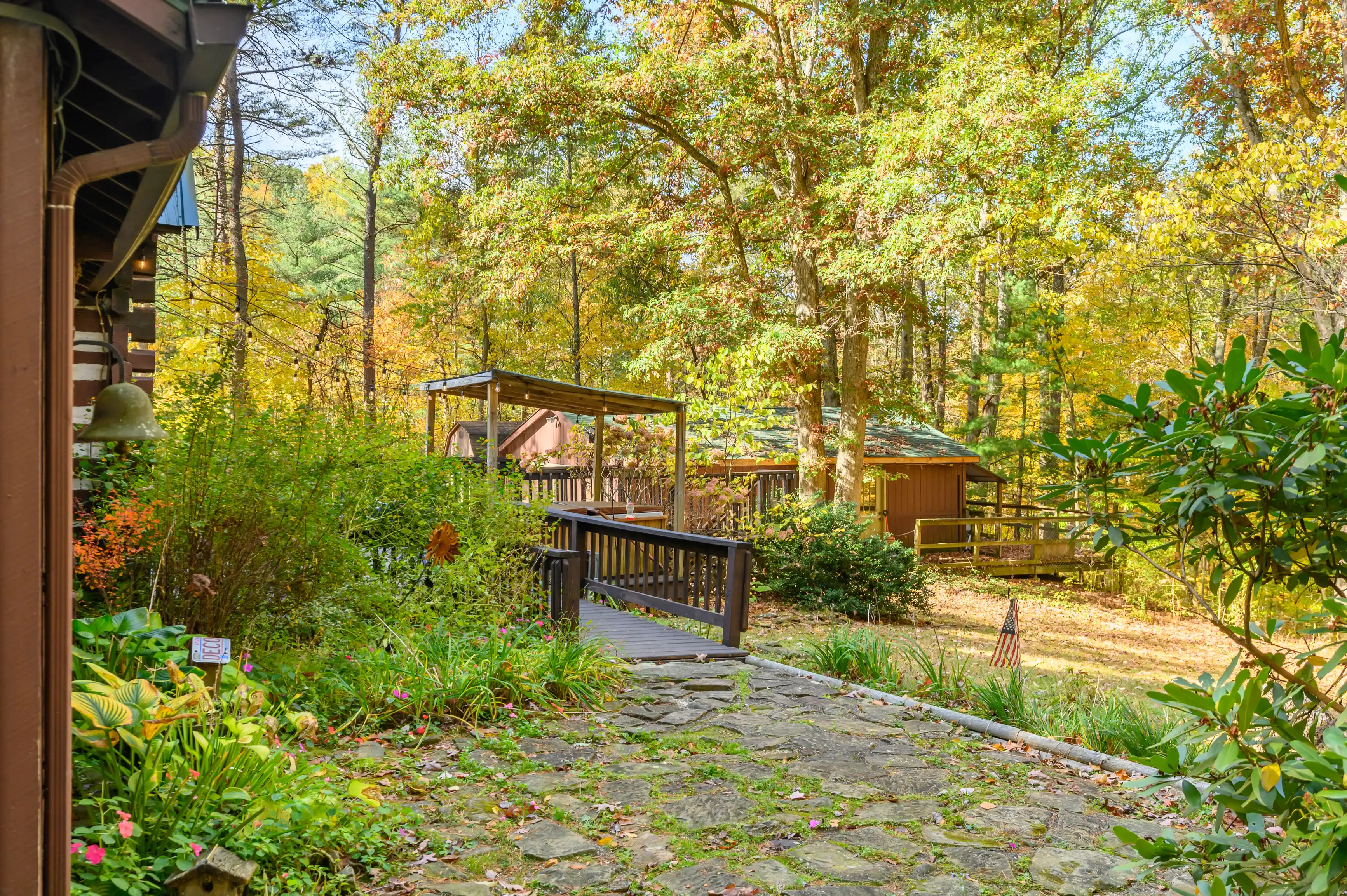 Stone pathway leading to a wooden bridge in a serene autumn forest setting with a cabin in the background and colorful foliage around.