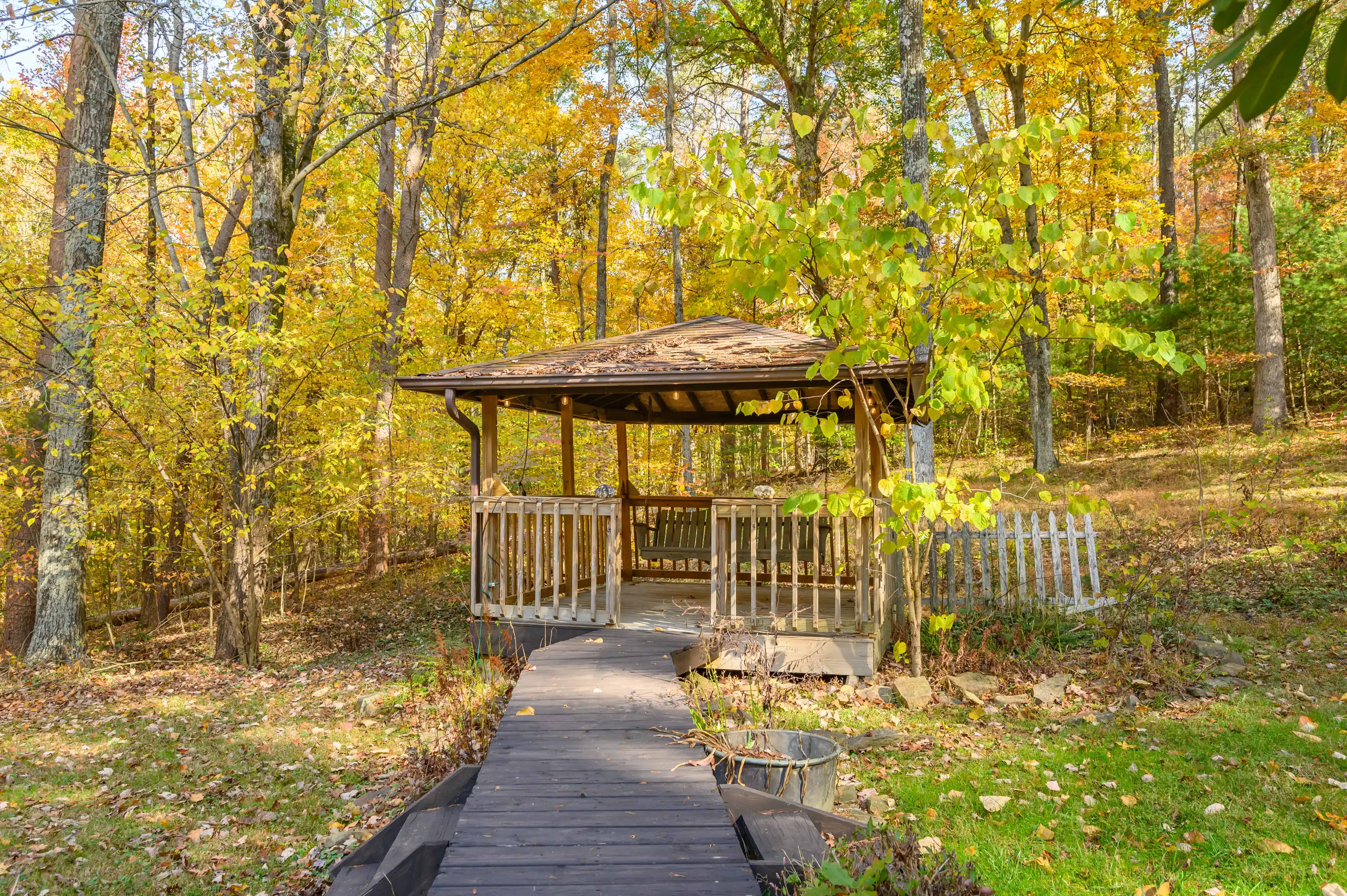 Wooden gazebo with a shingled roof at the end of a boardwalk surrounded by autumn-colored trees.