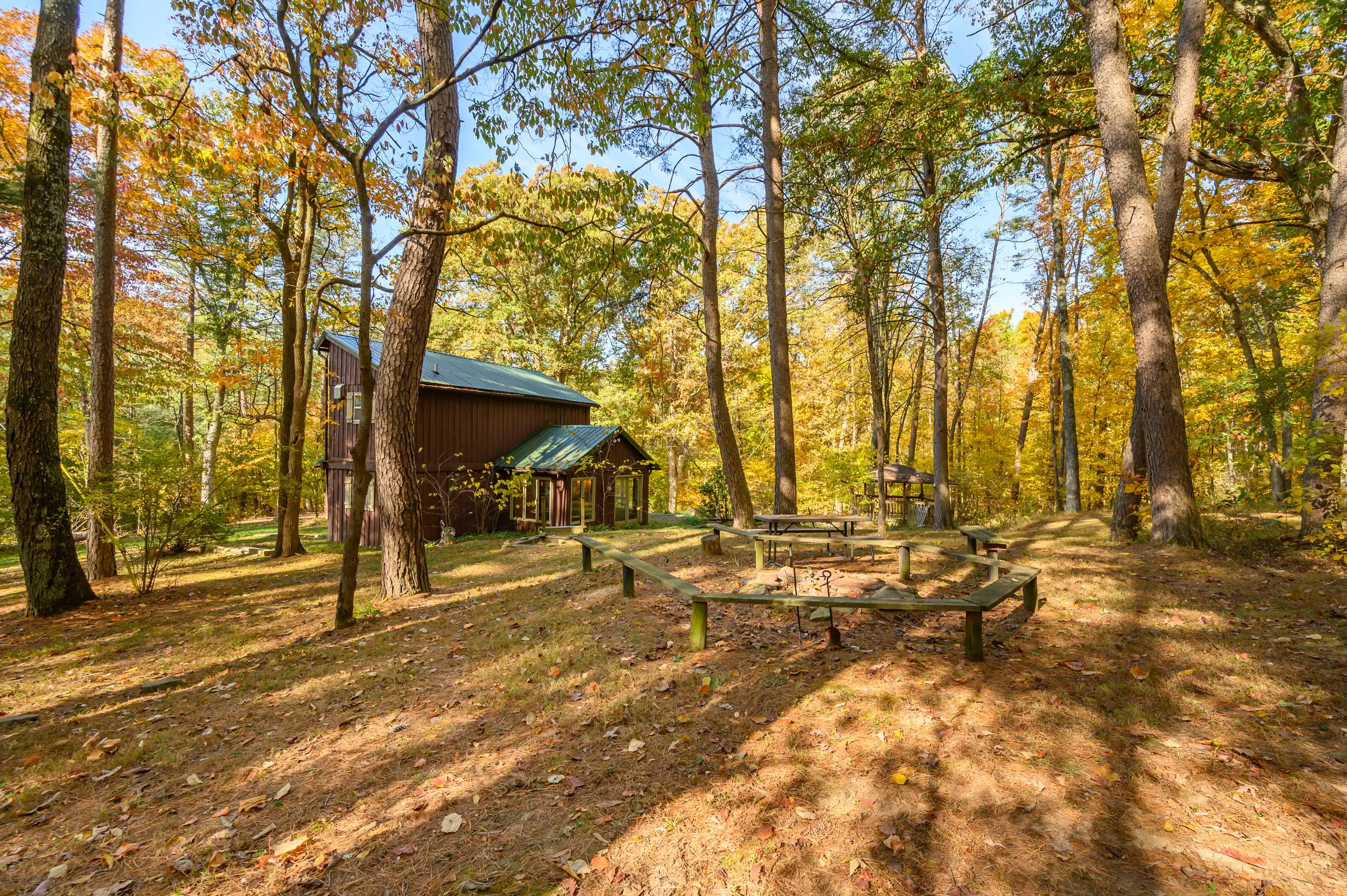 A peaceful woodland setting with a rustic cabin surrounded by tall trees with autumn foliage and a wooden picnic table in the foreground.