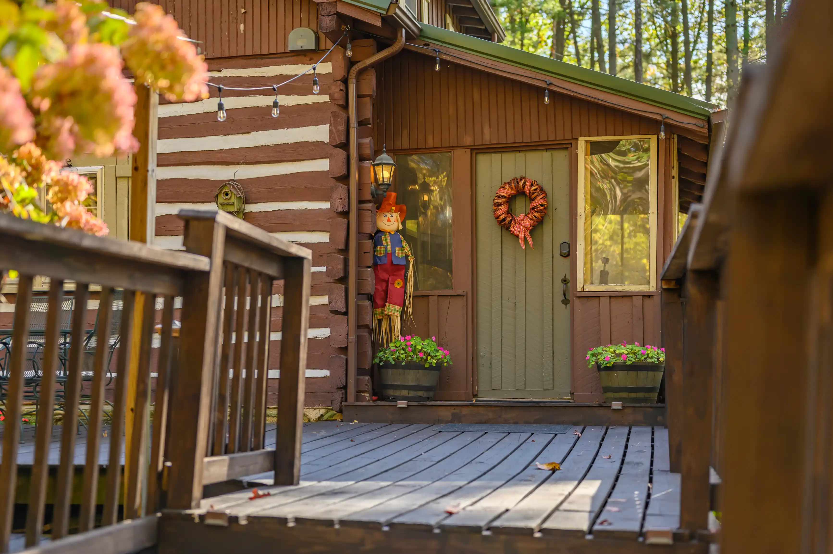 Cozy wooden cabin entrance with a heart-shaped wreath on the door, seasonal decorations, and surrounded by autumn foliage.